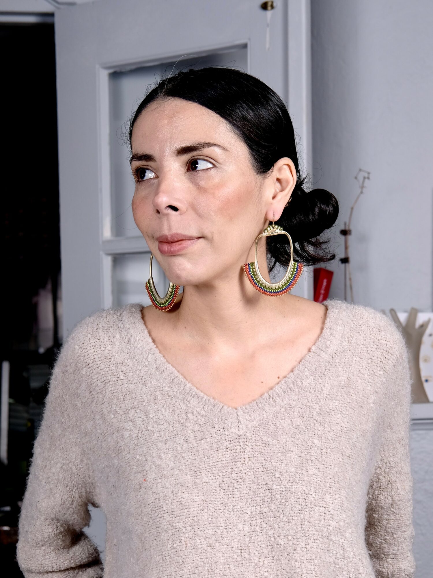 Woman with dark hair in a bun, wearing large earrings and a beige sweater, looking to the side.