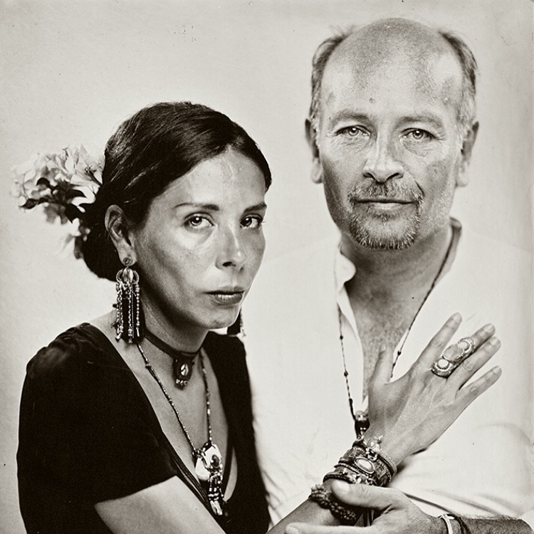 Two people, a woman and a man, pose closely together, with the woman touching the man's chest, both looking at the camera.
