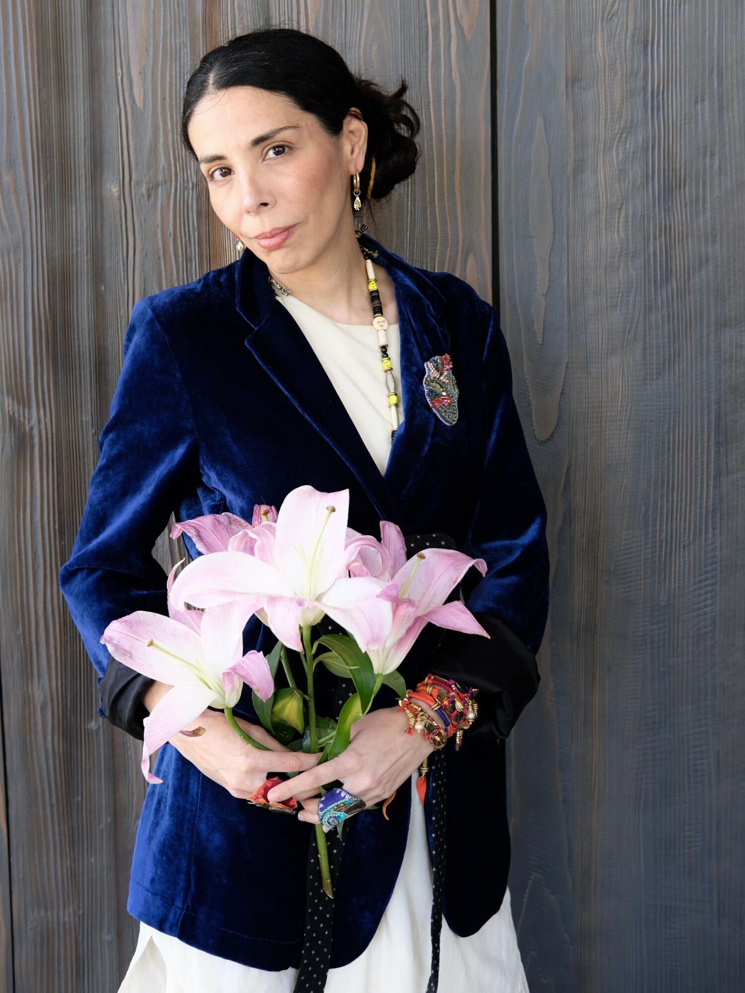 Woman with dark hair in a bun holding pink lilies, wearing a navy blue velvet blazer and jewelry.
