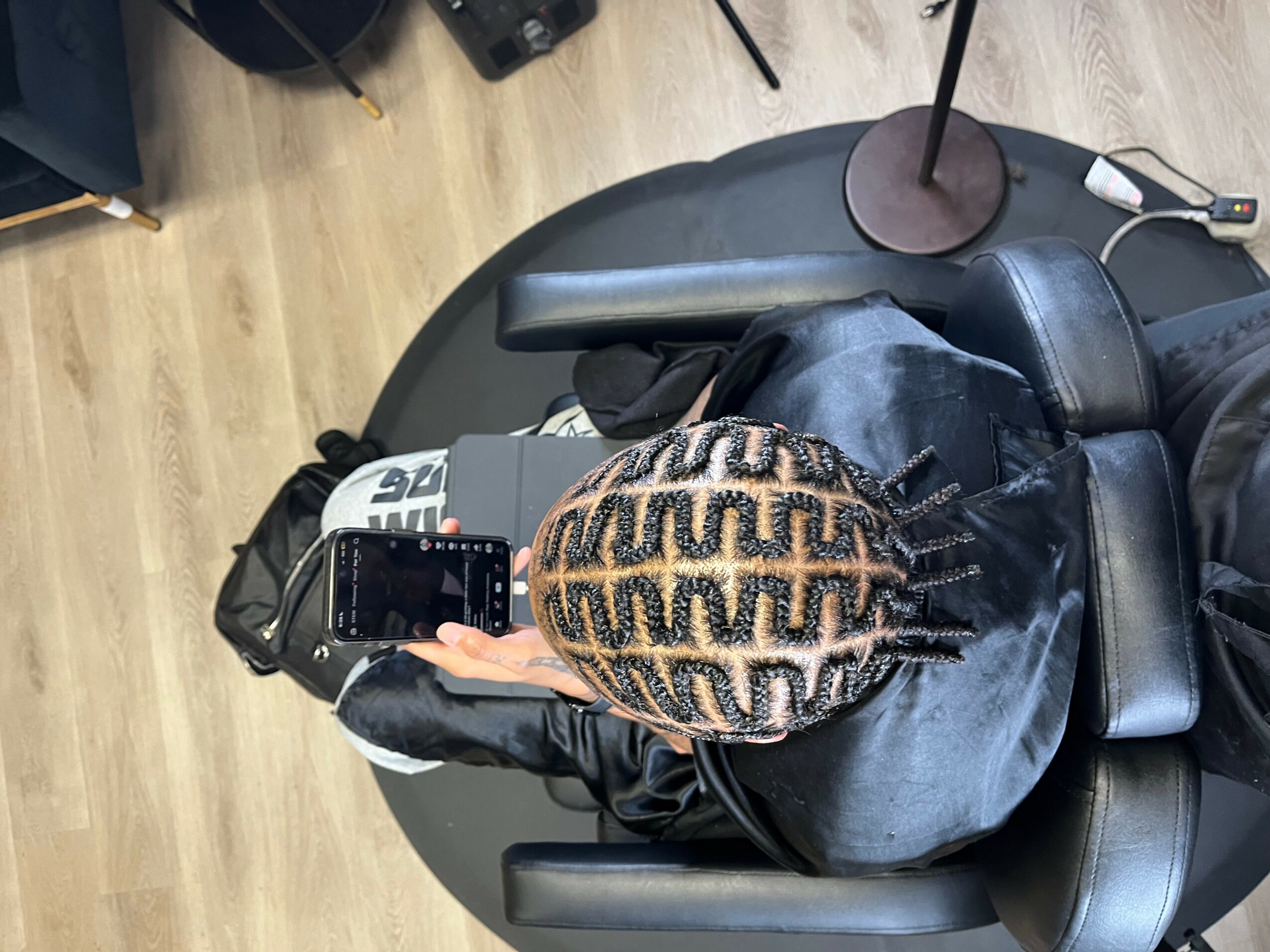 Top view of a person with braided hair sitting in a black chair, using a smartphone, in a room with a wooden floor.