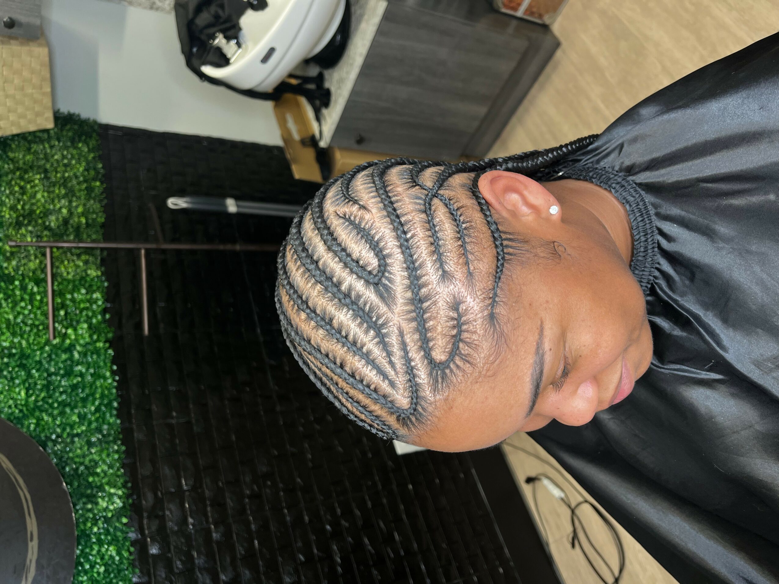 Side view of a person with intricate cornrow hairstyle, wearing a black cape, in a salon setting.