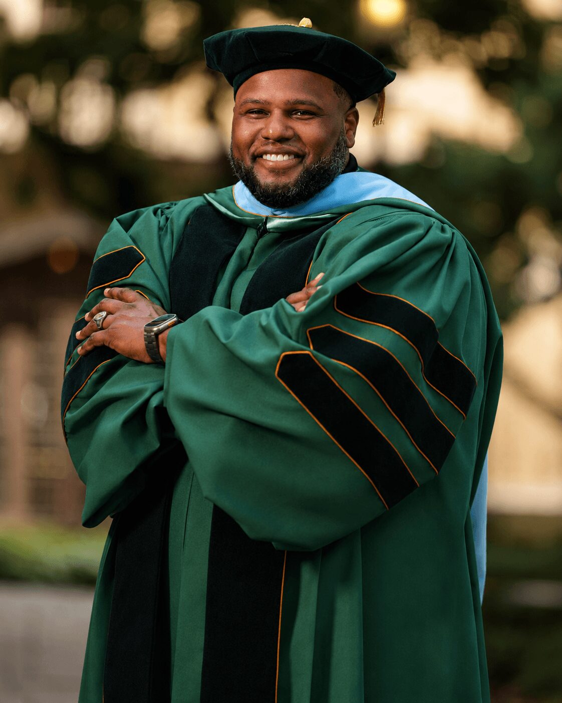 Man in graduation gown and cap smiling outdoors with arms crossed, trees in background.