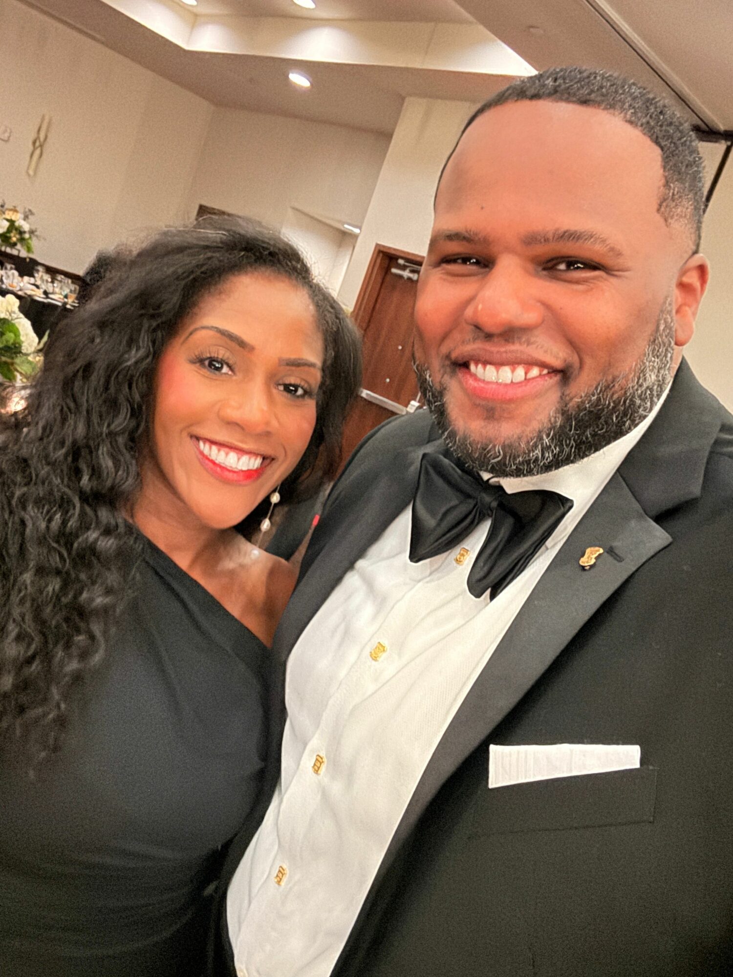 A smiling man in a tuxedo and a woman in a black dress pose together indoors.