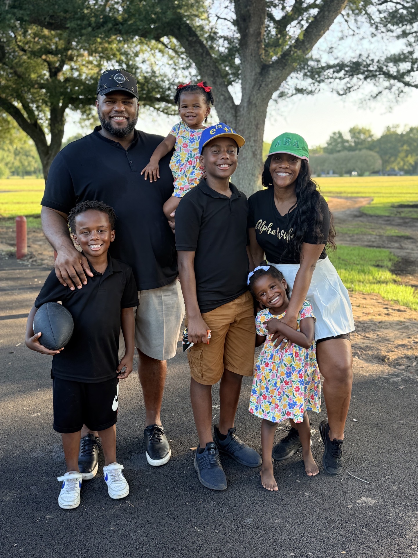 Family of six outdoors with trees and grass, smiling, standing on a paved path, children holding a ball and wearing colorful clothing.