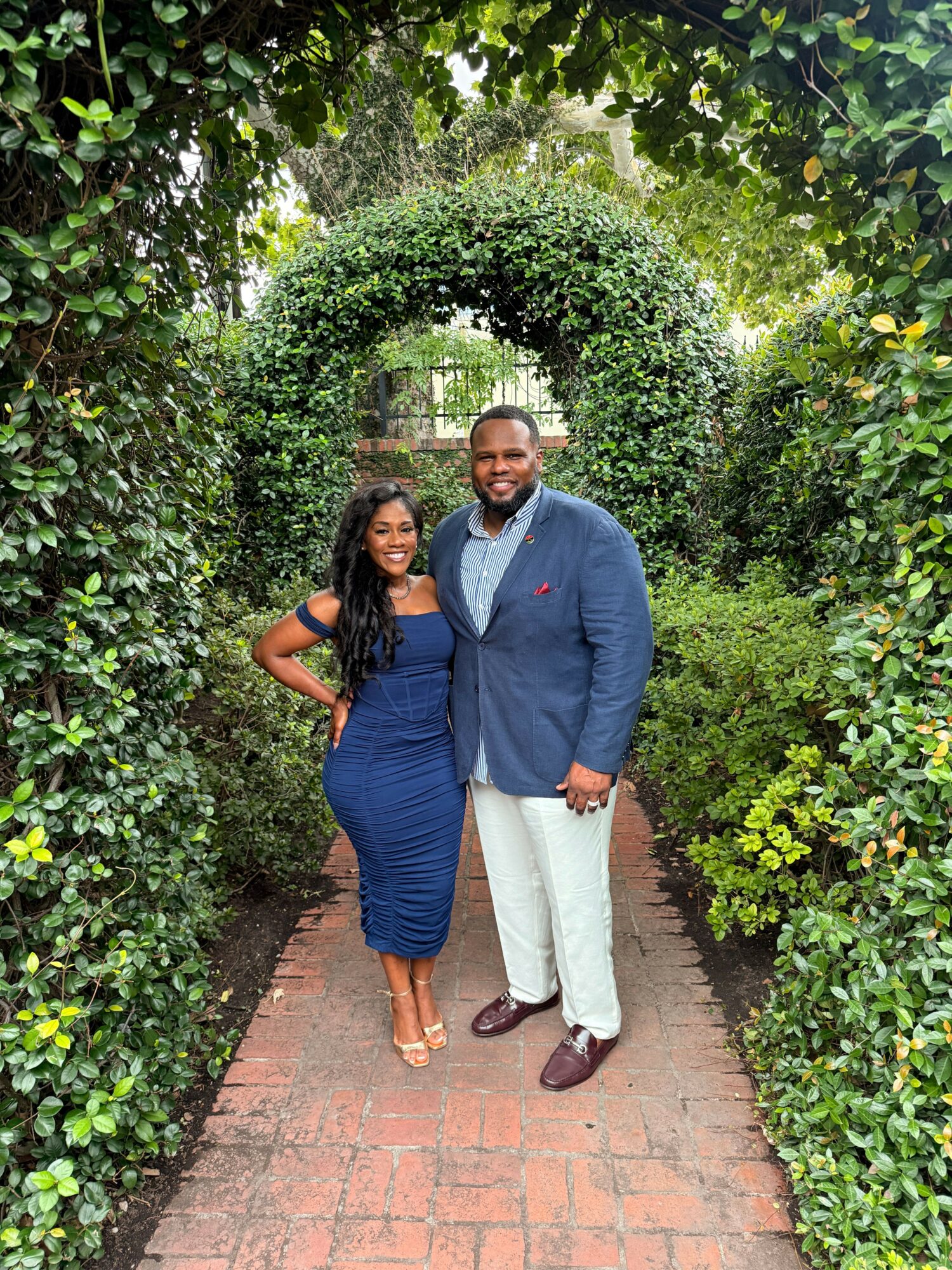 Two people standing on a brick path surrounded by green bushes and trees, smiling at the camera.