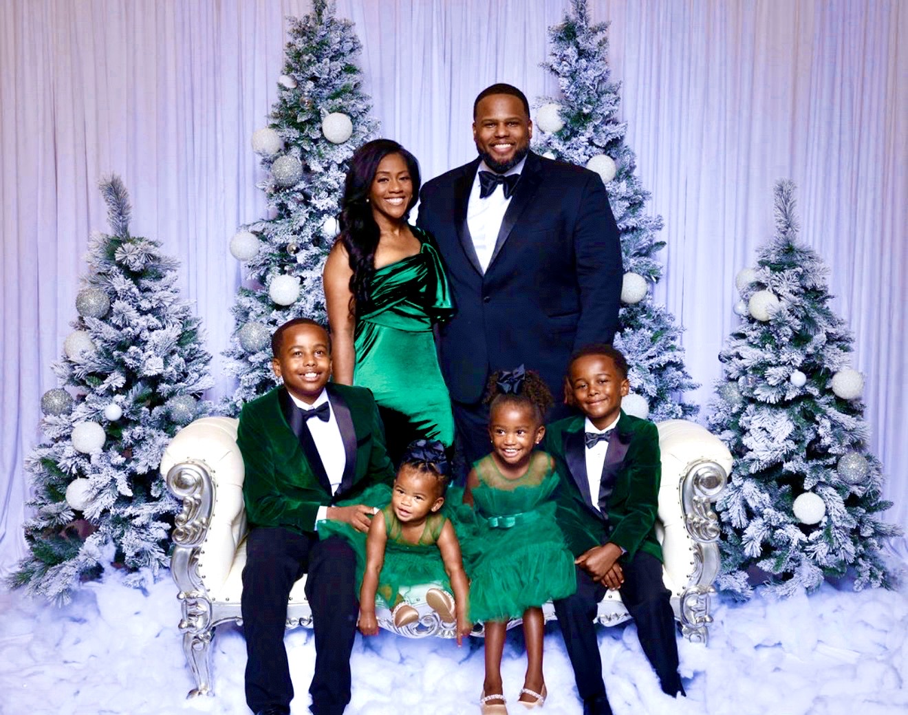 Family of six dressed in formal attire, standing in front of snow-covered Christmas trees, smiling happily.