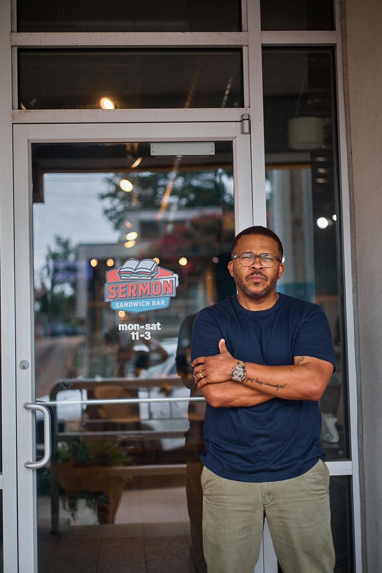 Man with glasses and beard stands outside a glass door, arms crossed, wearing a navy shirt and light pants.