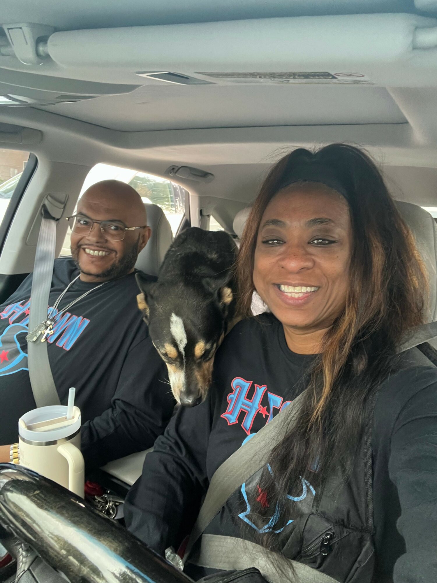 Two women and a small dog inside a car, smiling at the camera, with a drink cup on the dashboard.