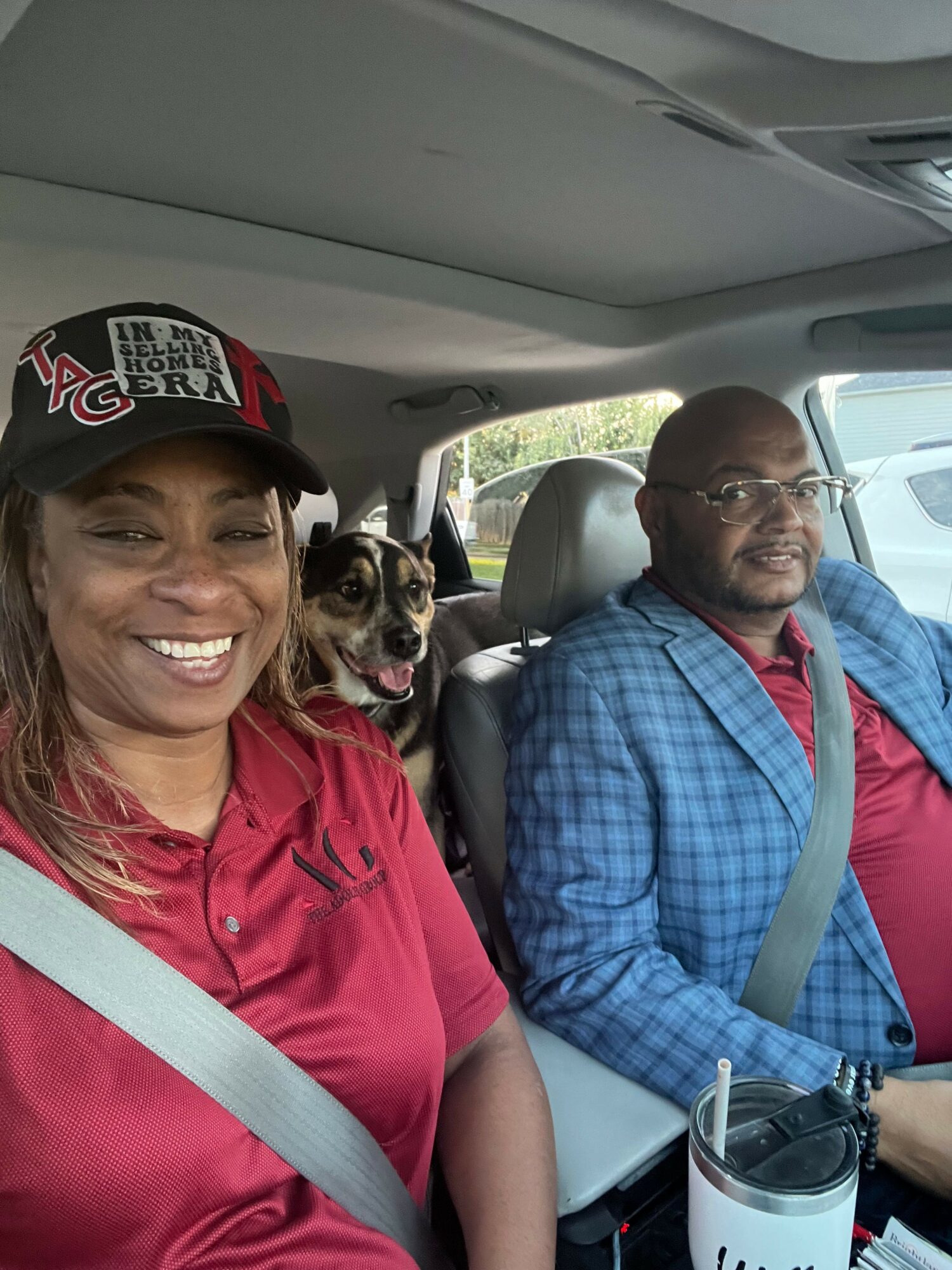 Two people and a dog sitting in a car, smiling, with the woman in the front seat and the man in the back seat.