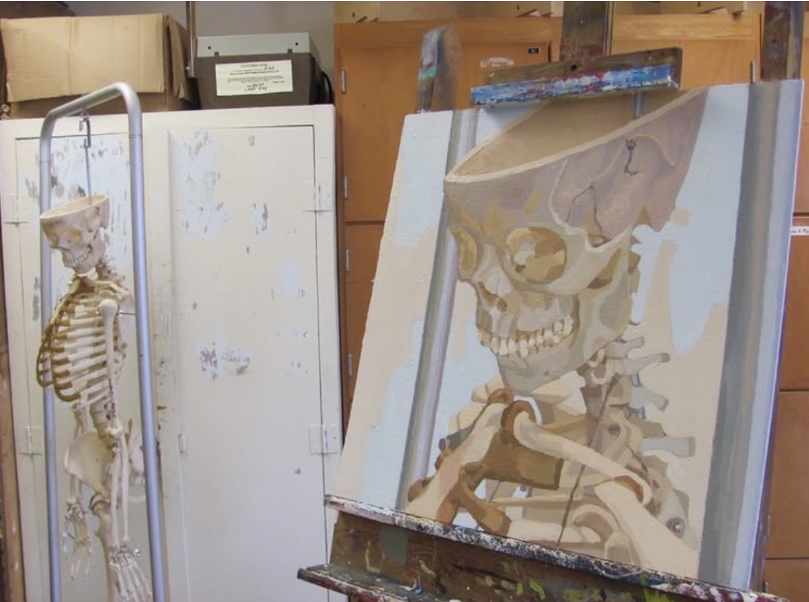 A skeleton model stands next to an easel with a painting of a skull and bones, in an art studio.
