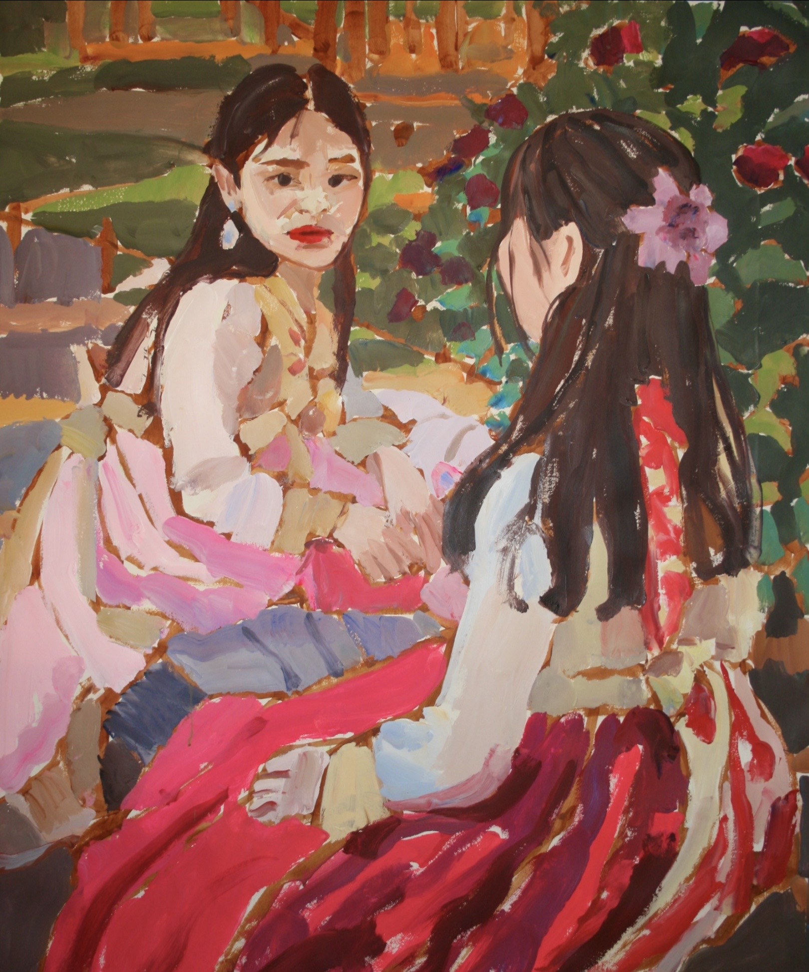 Two girls sitting outdoors, one with long dark hair and a pink flower in her hair, the other with long dark hair, wearing colorful clothing, engaged in conversation.