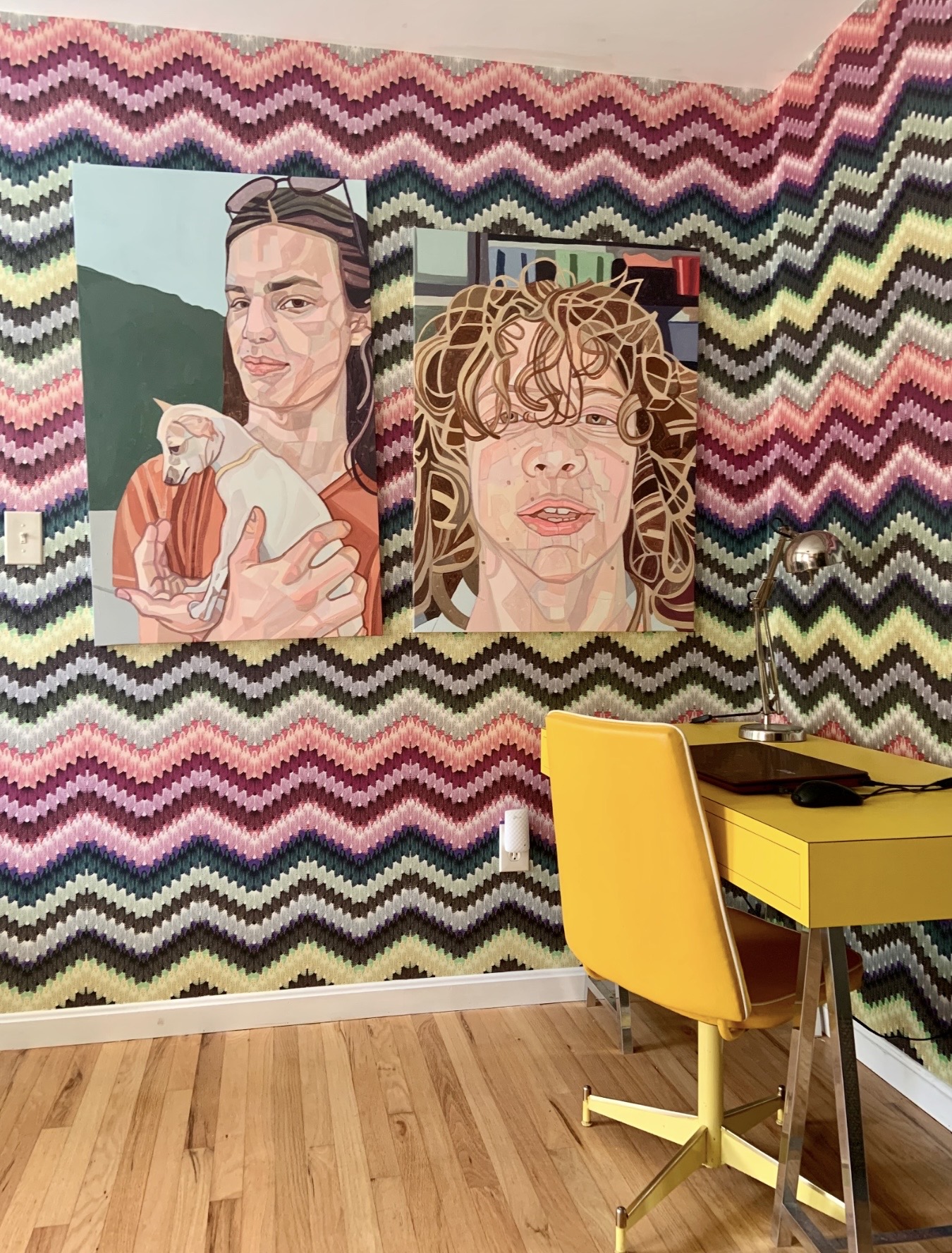 Two large portrait paintings on a zigzag patterned wall, one of a woman holding a cat, the other of a woman with curly hair, yellow chair and desk in foreground.