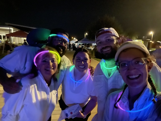 Group of five people smiling at night, wearing glow necklaces and headbands, outdoors with lights and tents in background.