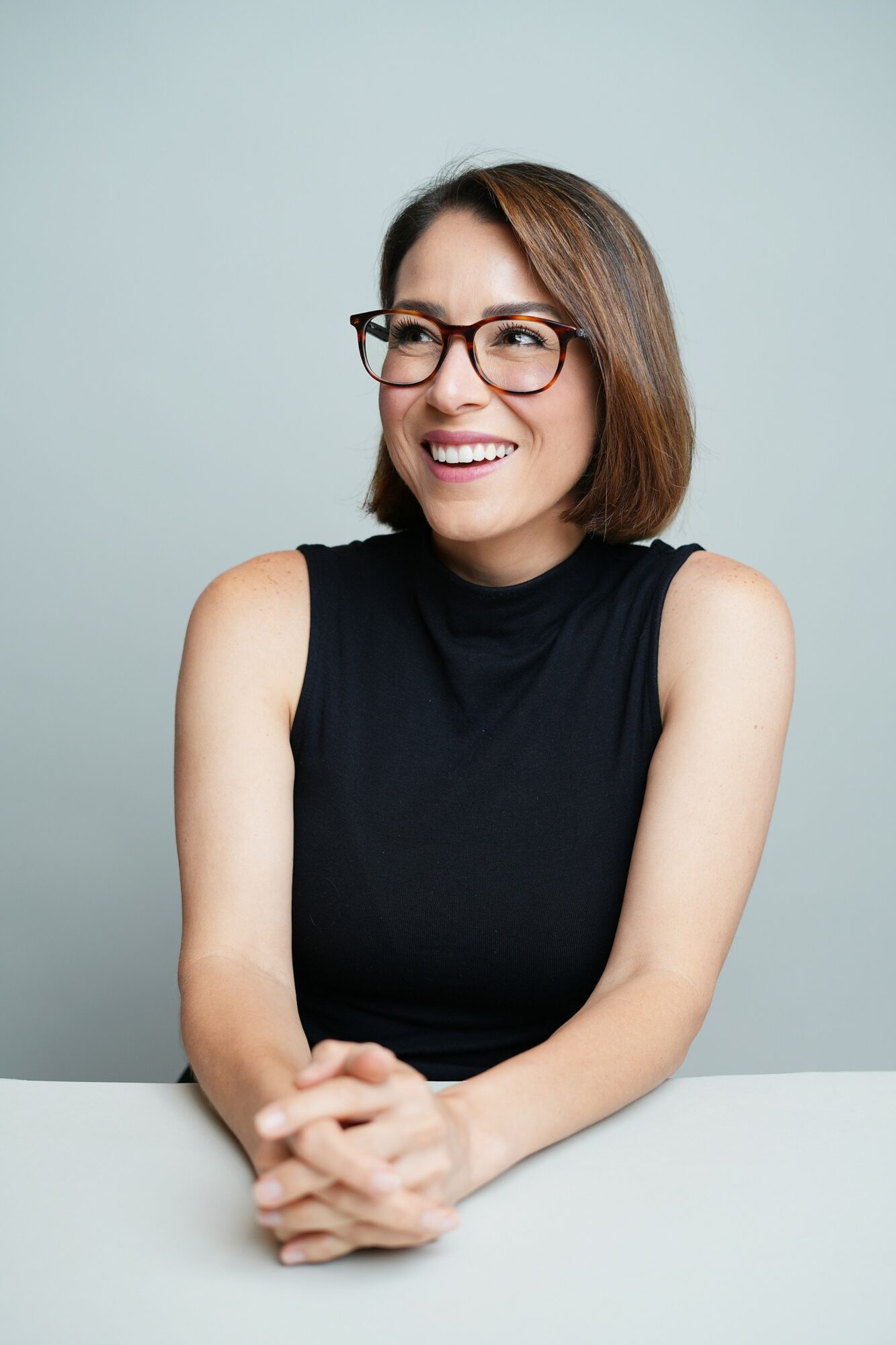 Woman with glasses smiling, wearing a sleeveless black top, sitting with hands clasped, against a light blue background.