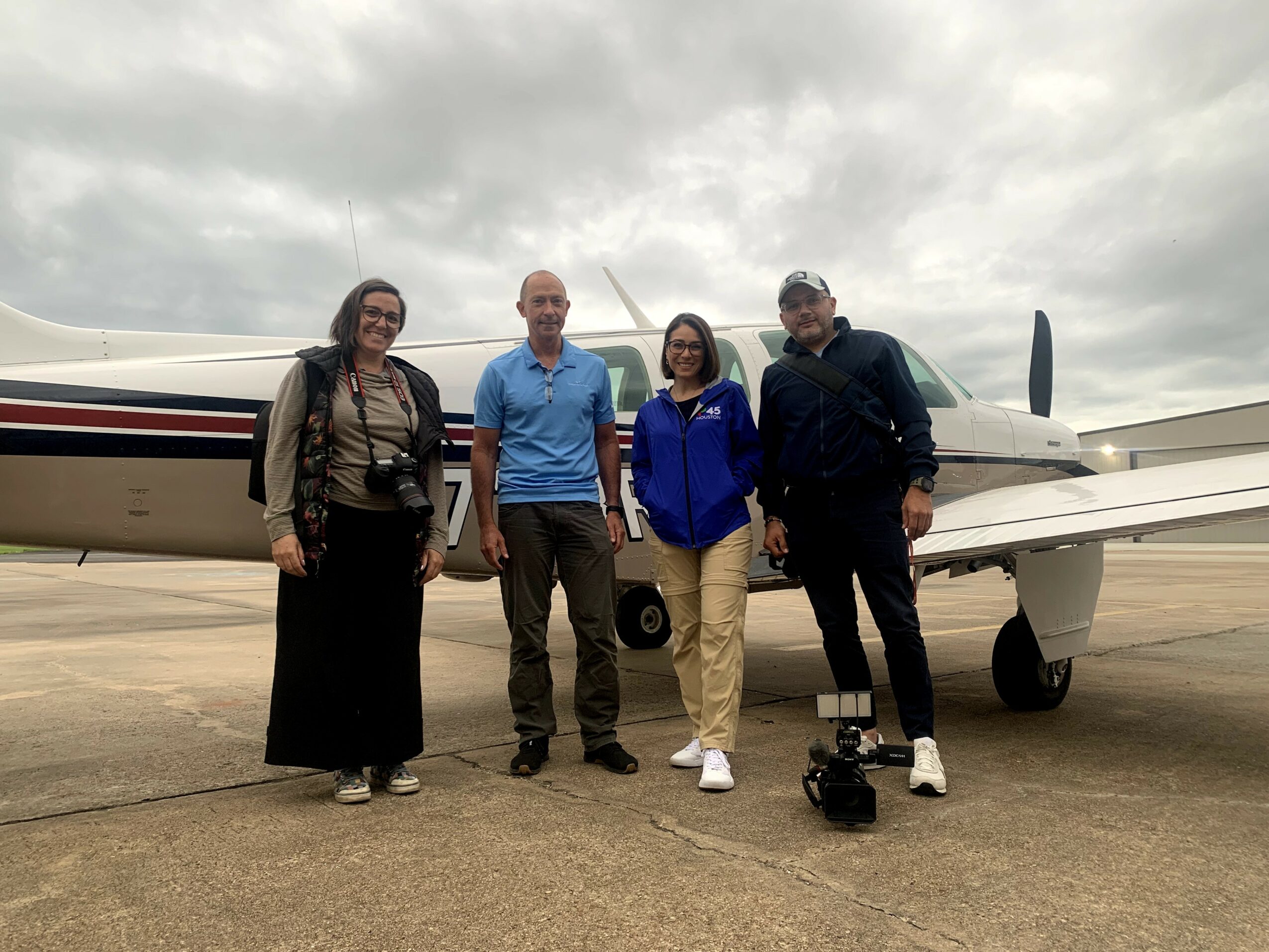 Four people stand in front of a small airplane on a cloudy day, smiling and posing for the photo.