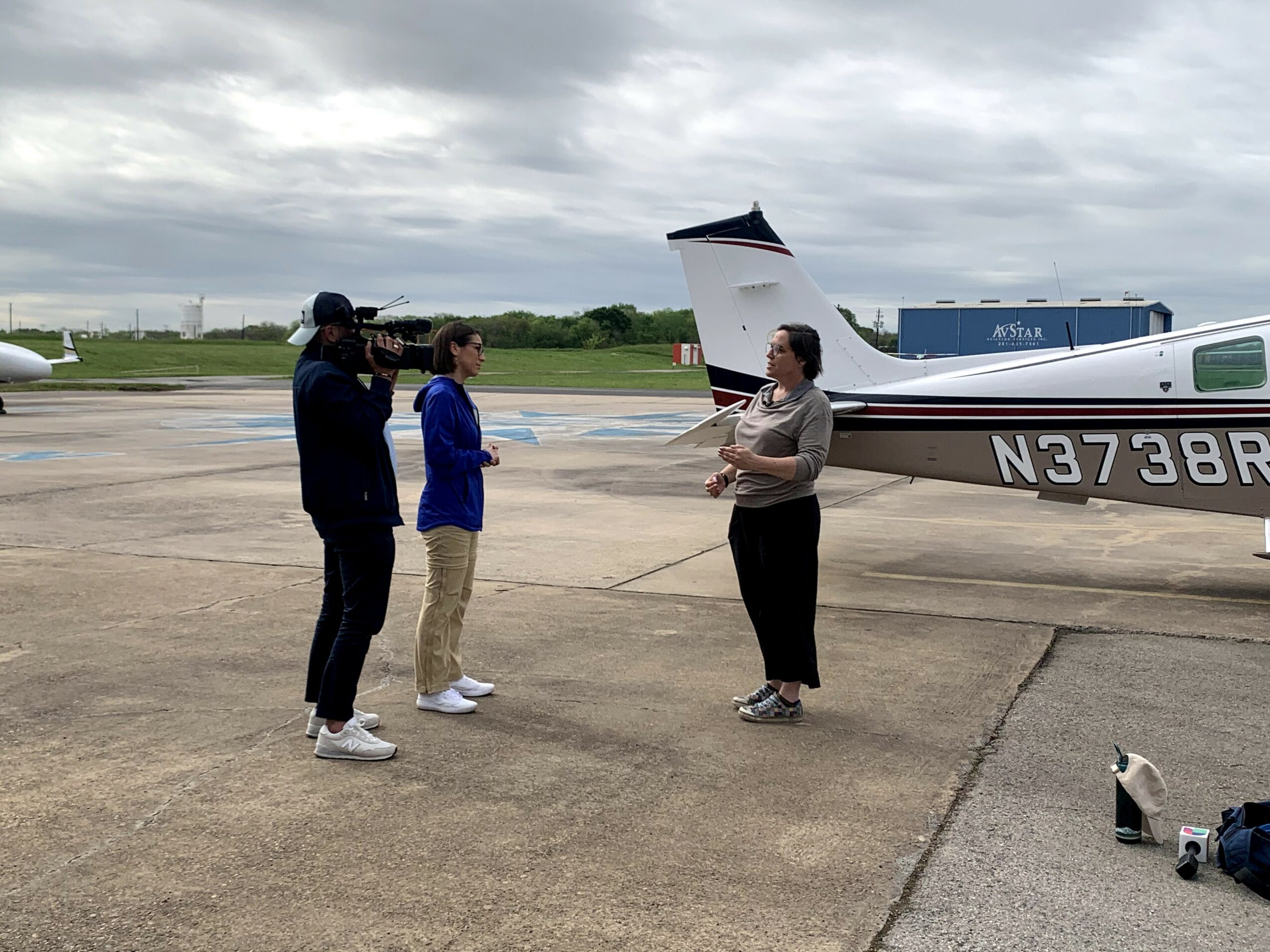Three people standing on an airport tarmac near small aircraft, engaged in conversation under cloudy sky.