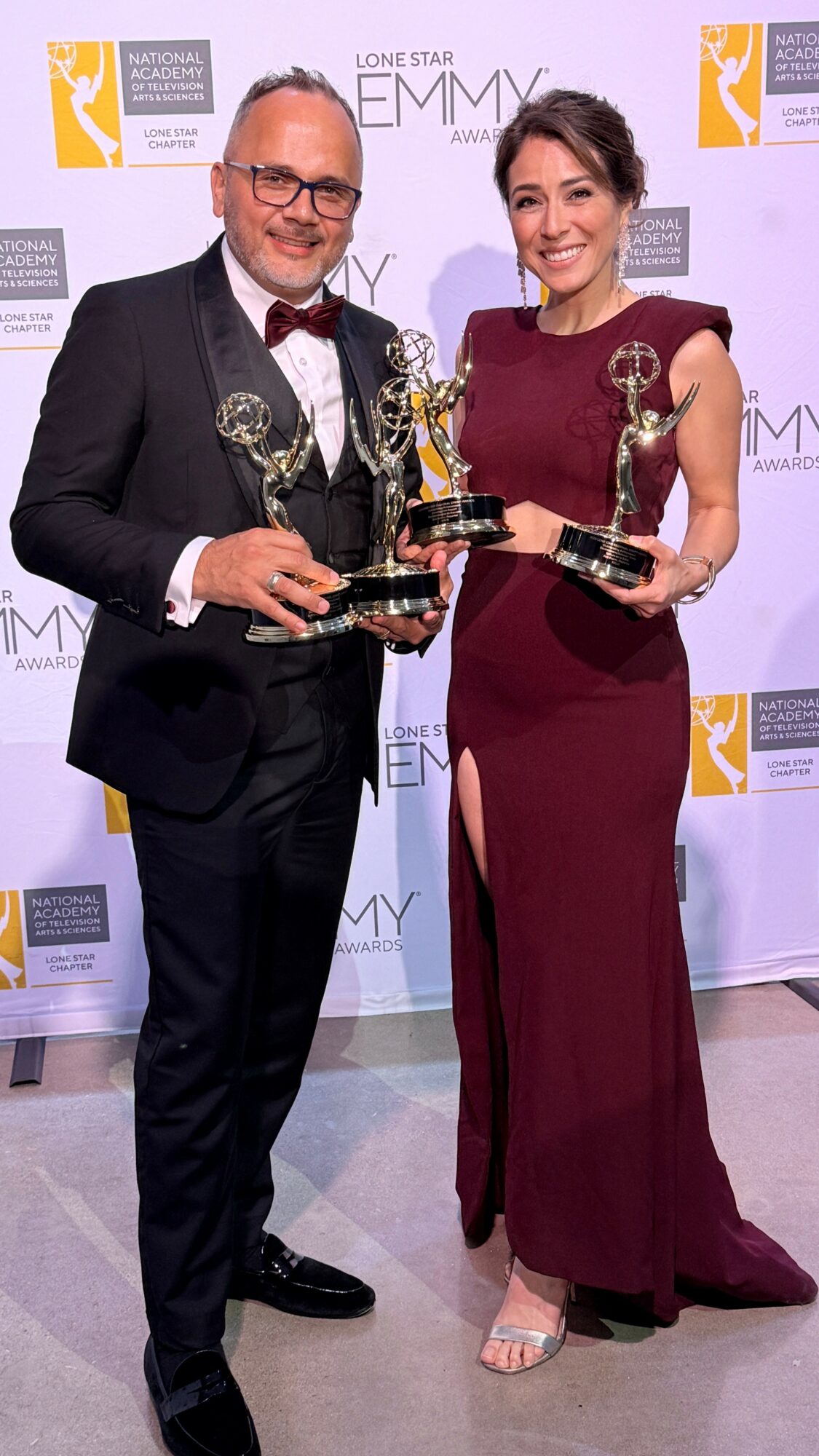 Man and woman holding Emmy awards, smiling, standing in front of a backdrop with logos, dressed formally.
