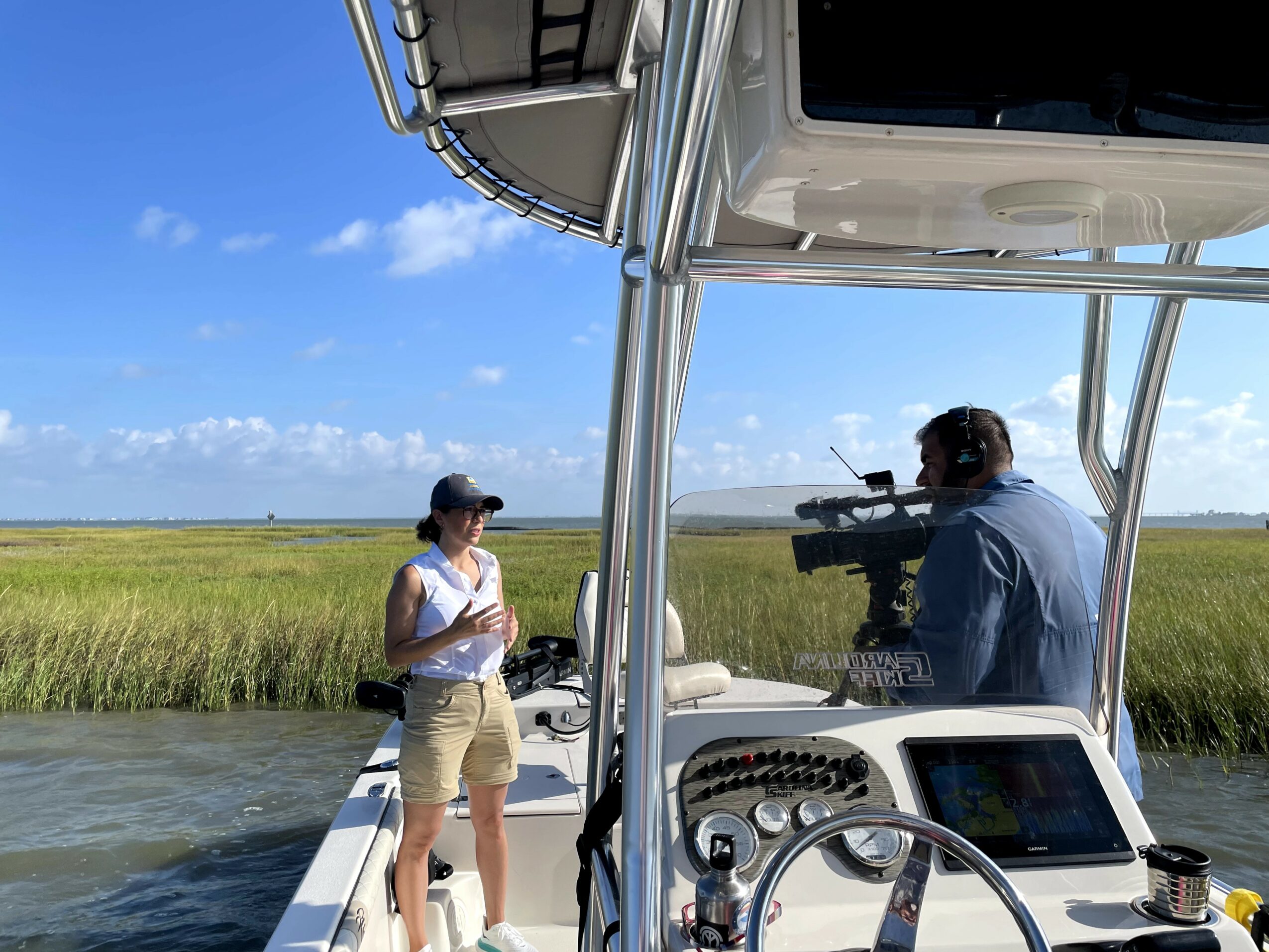 Two people on a boat in a marsh, one holding a camera and the other standing with a notepad, blue sky above.