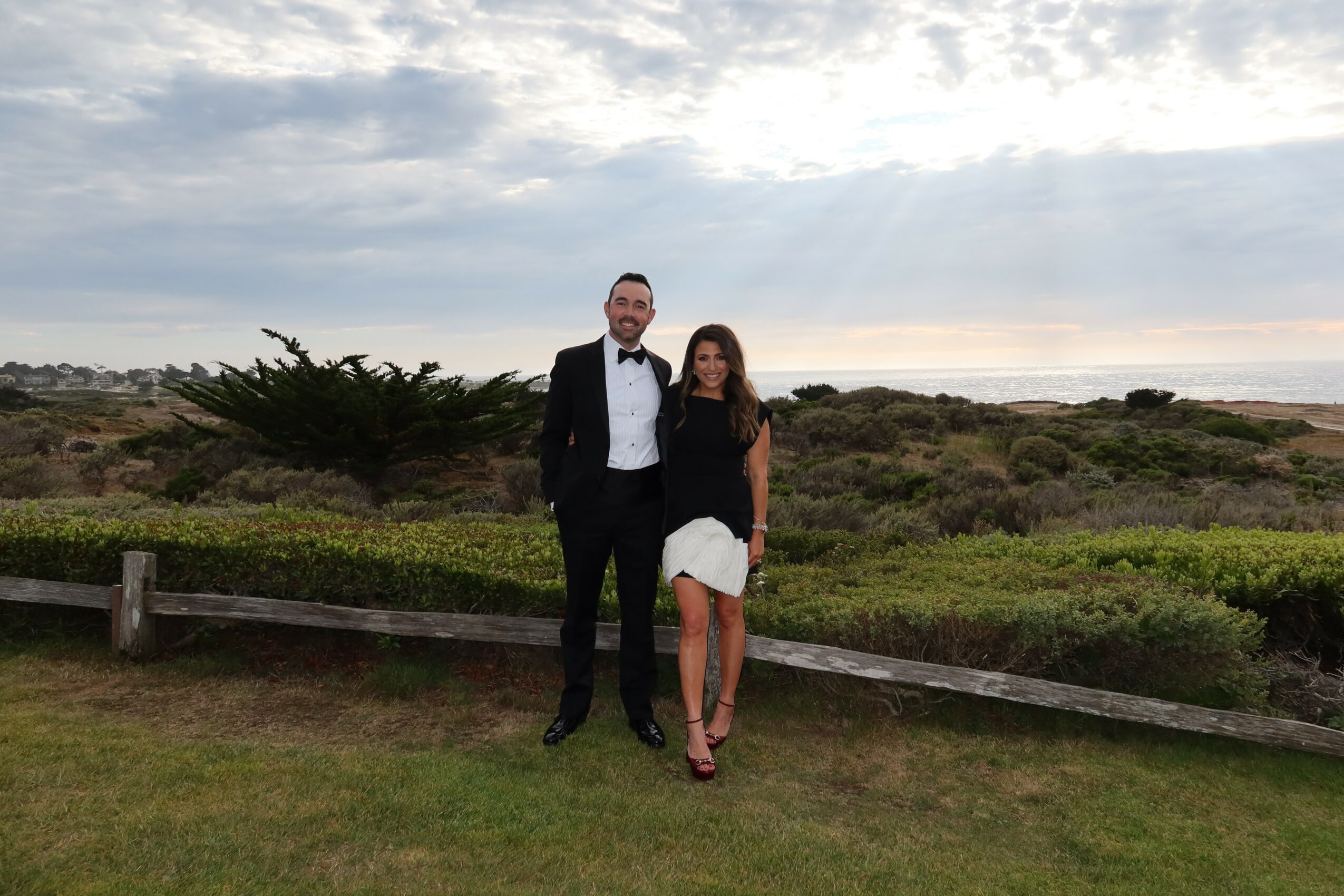A man in a tuxedo and a woman in a black top and white skirt stand outdoors on grass with a landscape and cloudy sky behind them.
