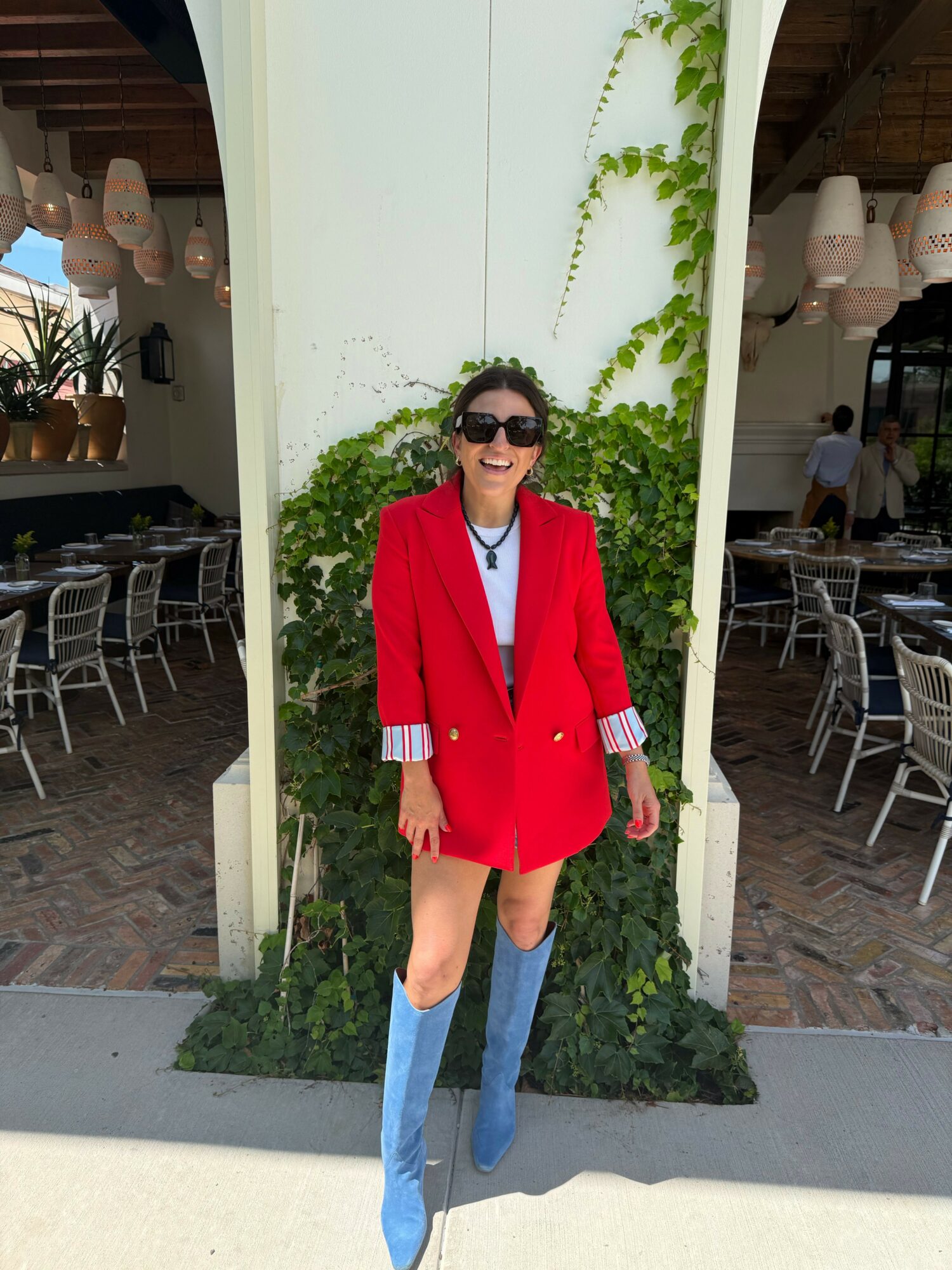 Person in red coat and sunglasses standing outside near a white wall and greenery.