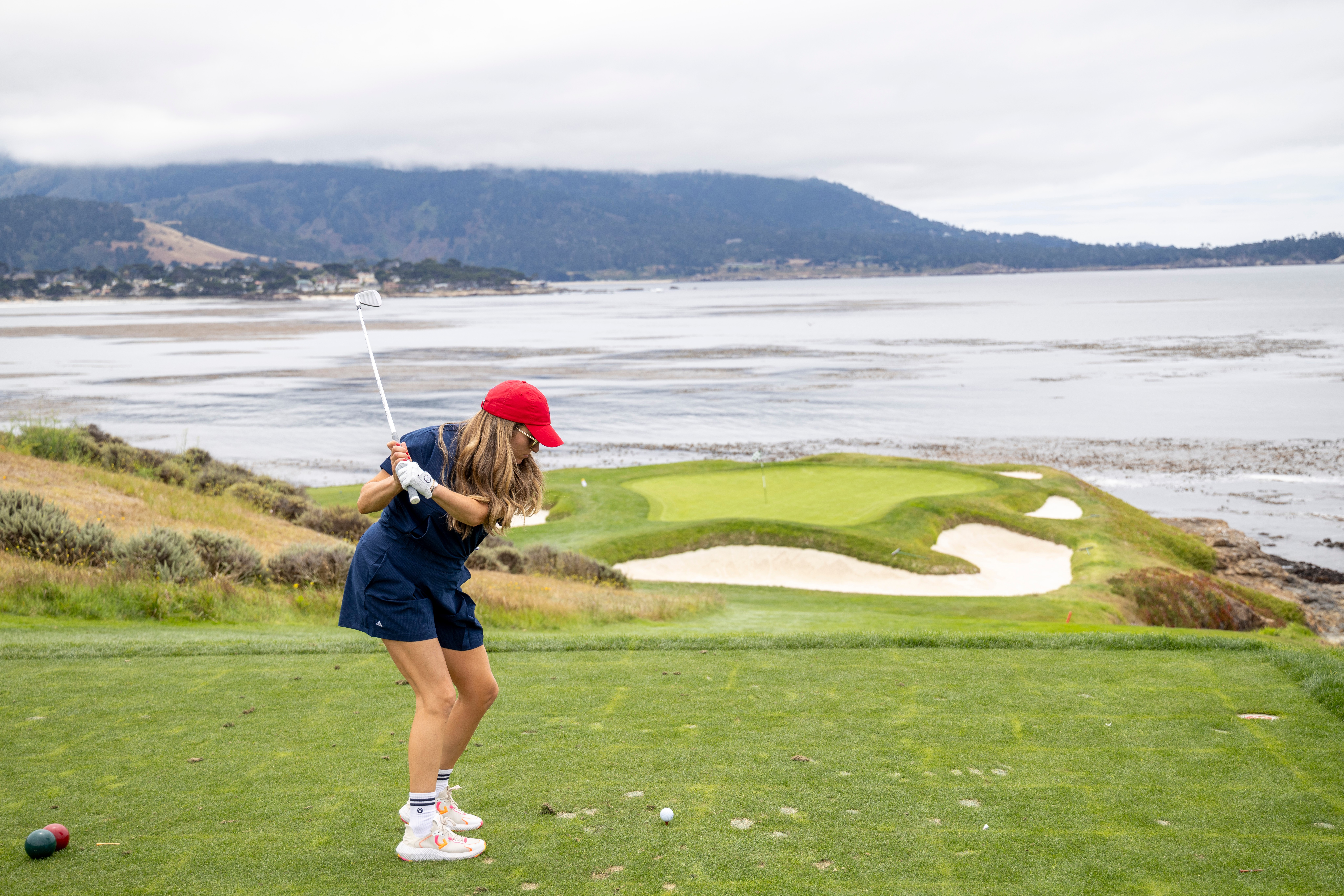 Girl in red cap and navy outfit swings golf club on green near water with hills in background.