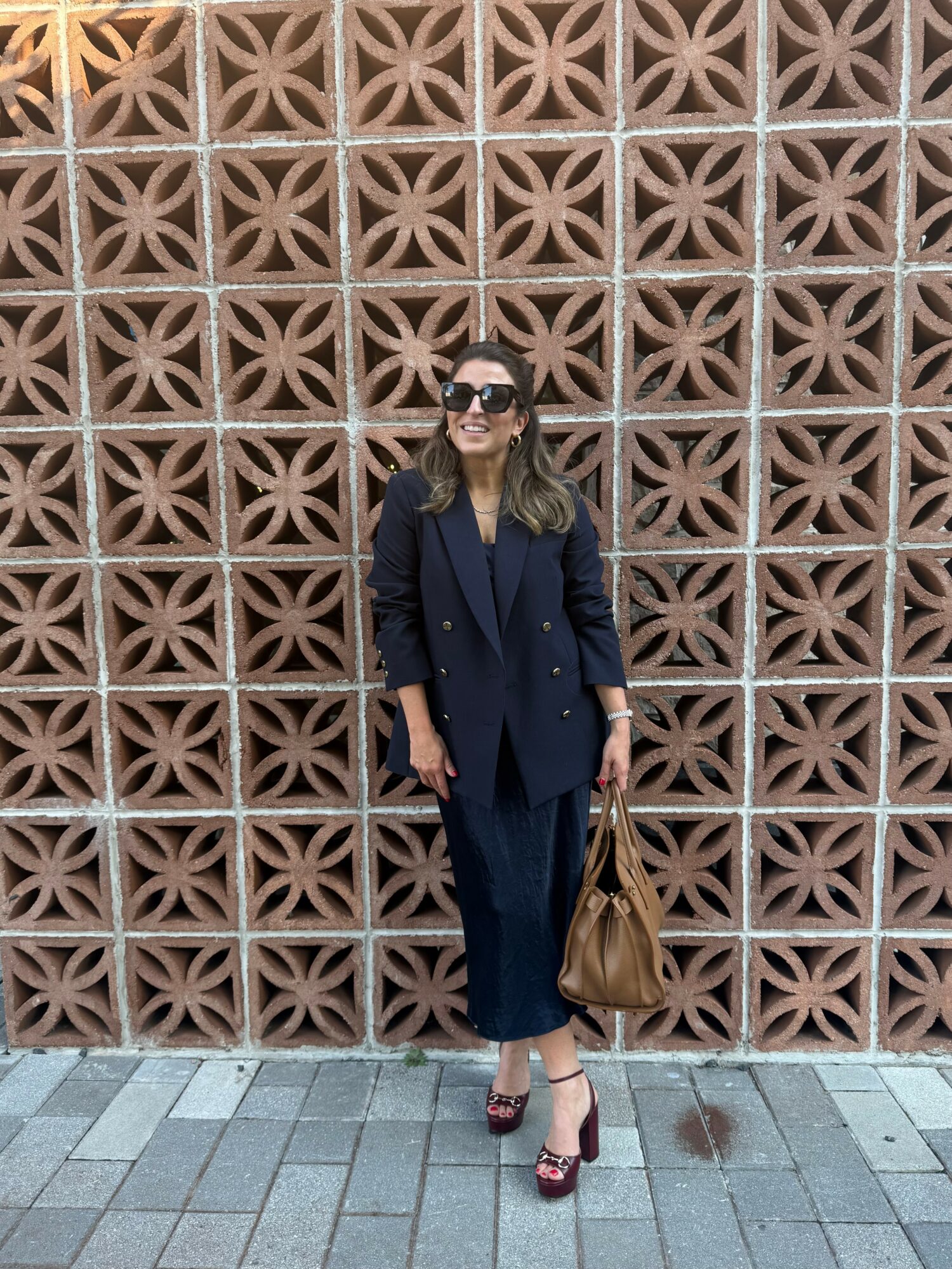 Woman in dark suit with sunglasses holding a brown bag standing in front of a decorative brick wall.