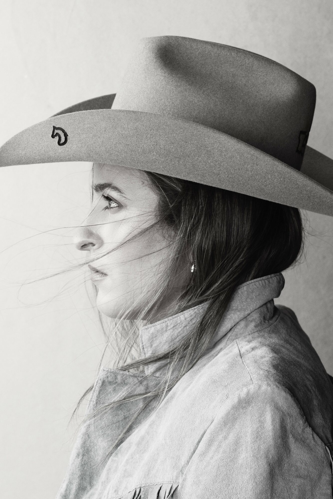 Side profile of a woman wearing a wide-brimmed hat and a jacket, with her hair flowing in front of her face.