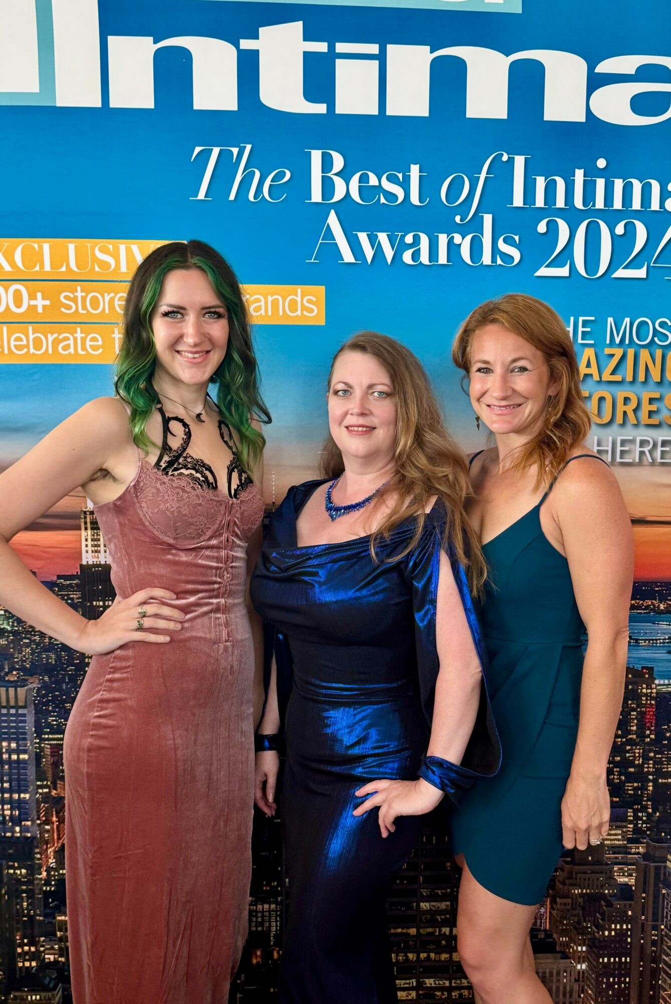Three women in elegant dresses standing together at an awards event, smiling, with a cityscape background and a blue banner above.