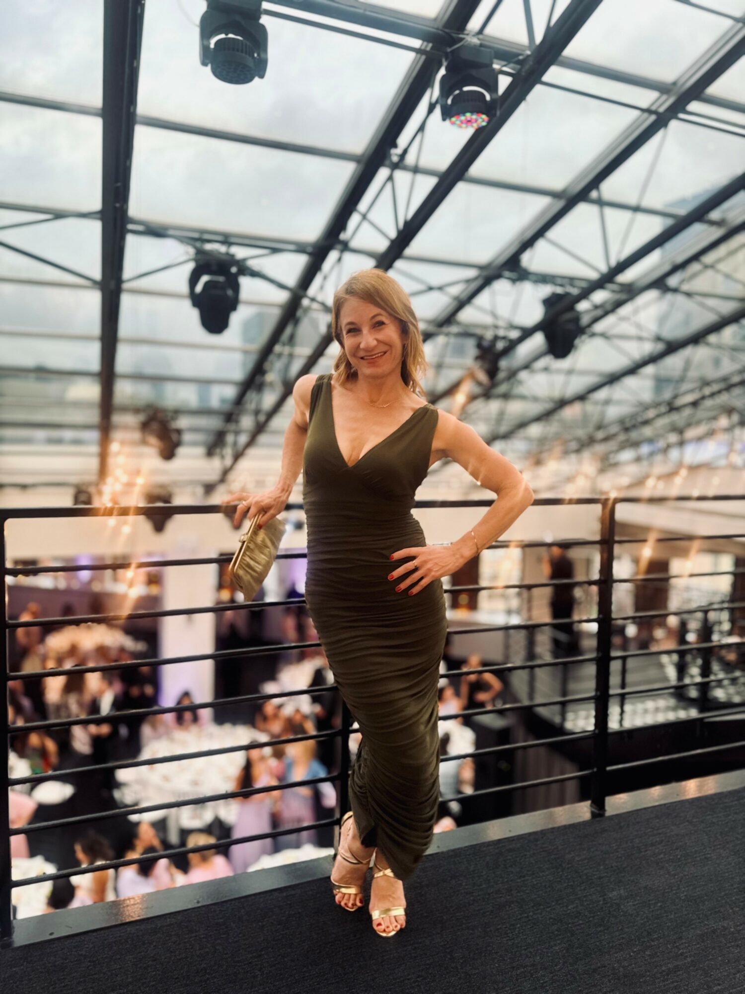 Woman in a black dress standing on a balcony with a glass ceiling, smiling, with a crowd below.