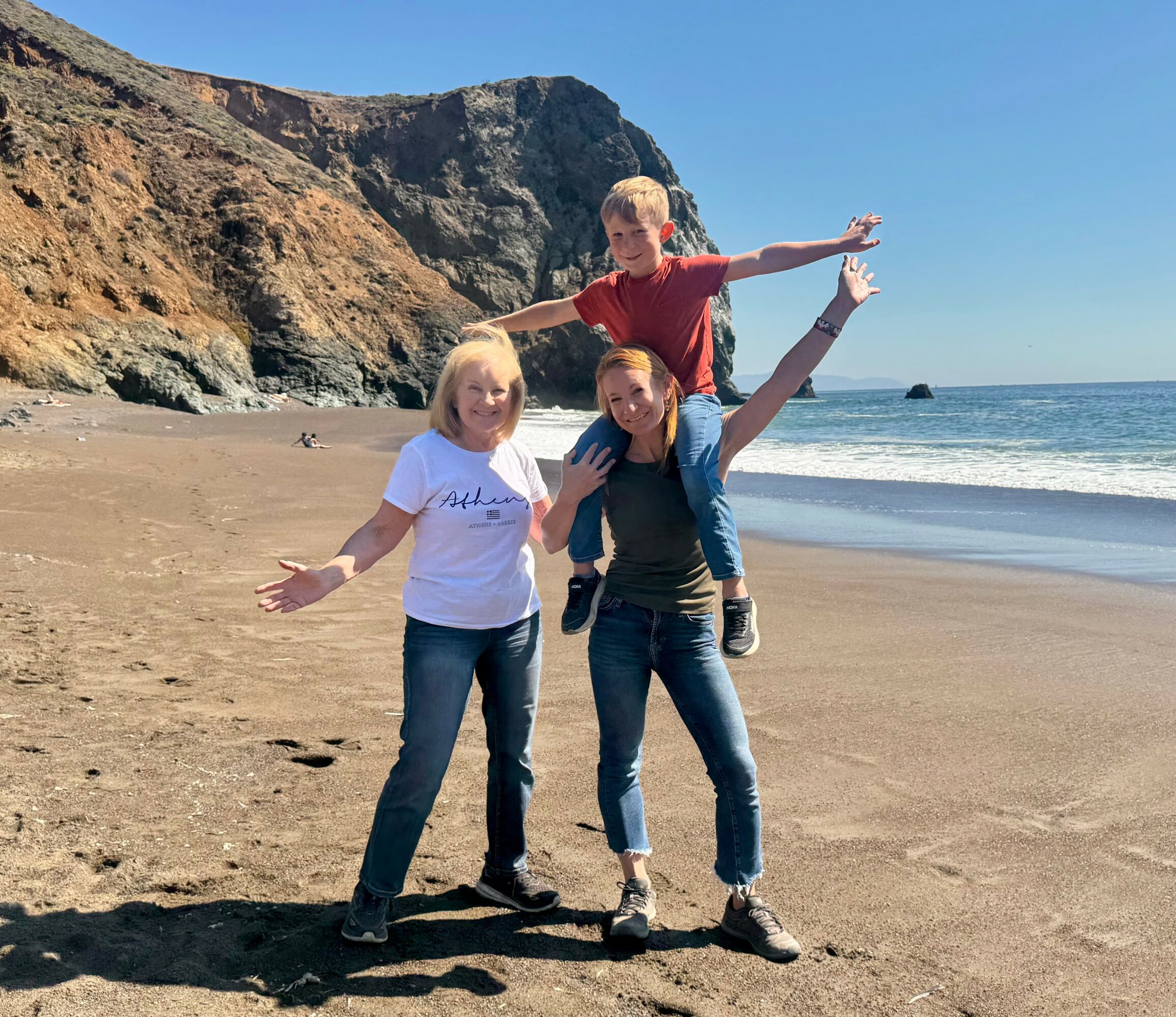 Three people on a beach with cliffs in the background, two women standing and a boy on their shoulders, all smiling.