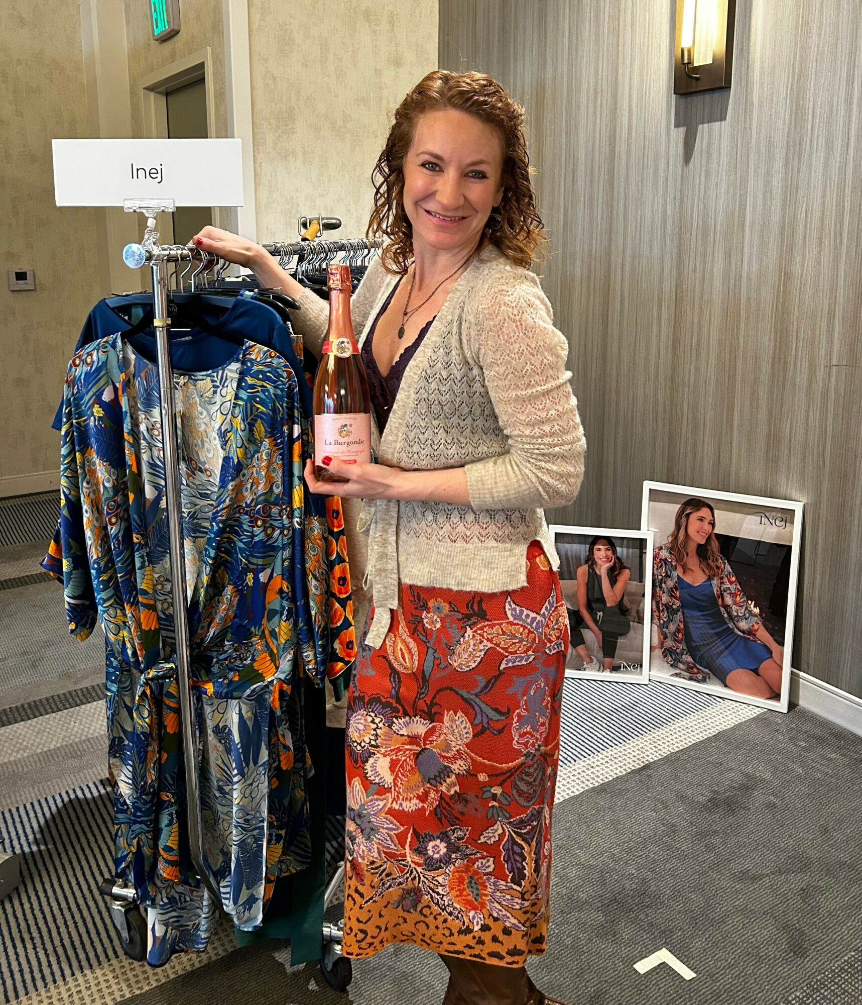 Woman with curly hair smiling, holding a pink bottle, standing next to a clothing rack with colorful dresses, in a room with framed photos on the floor.
