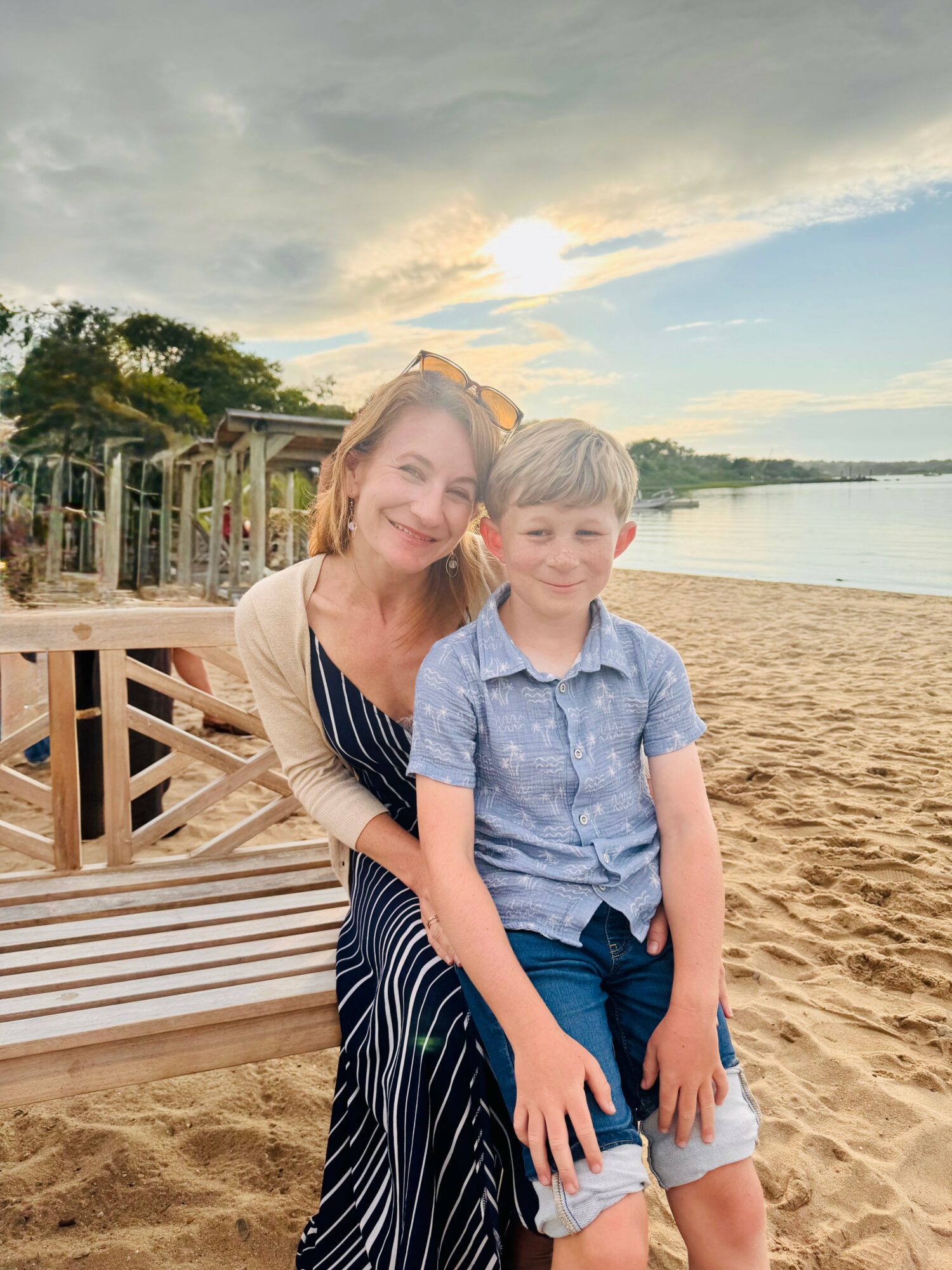 Woman and boy standing on sandy beach near water, smiling, with trees and sky in background.