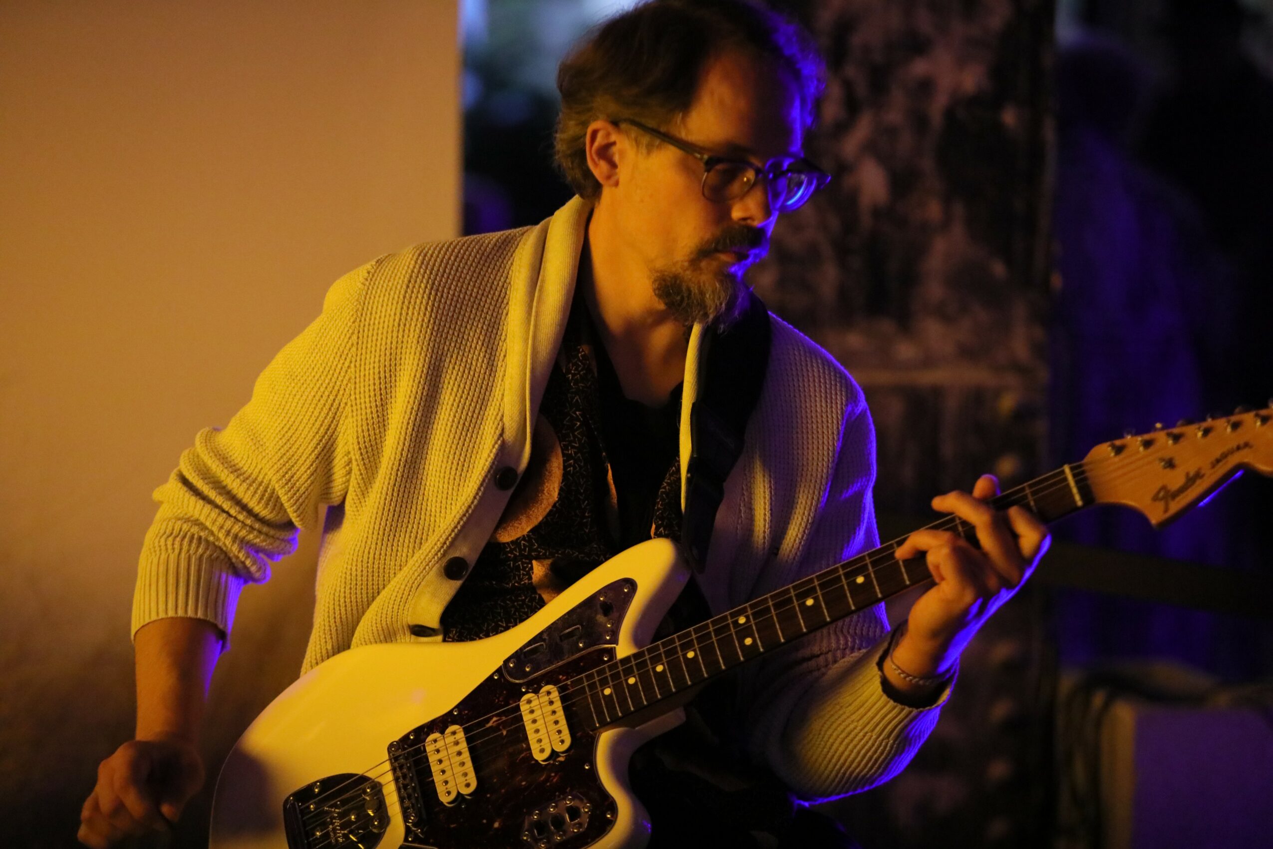 Person playing electric guitar, wearing glasses and a light-colored cardigan, in dim lighting.