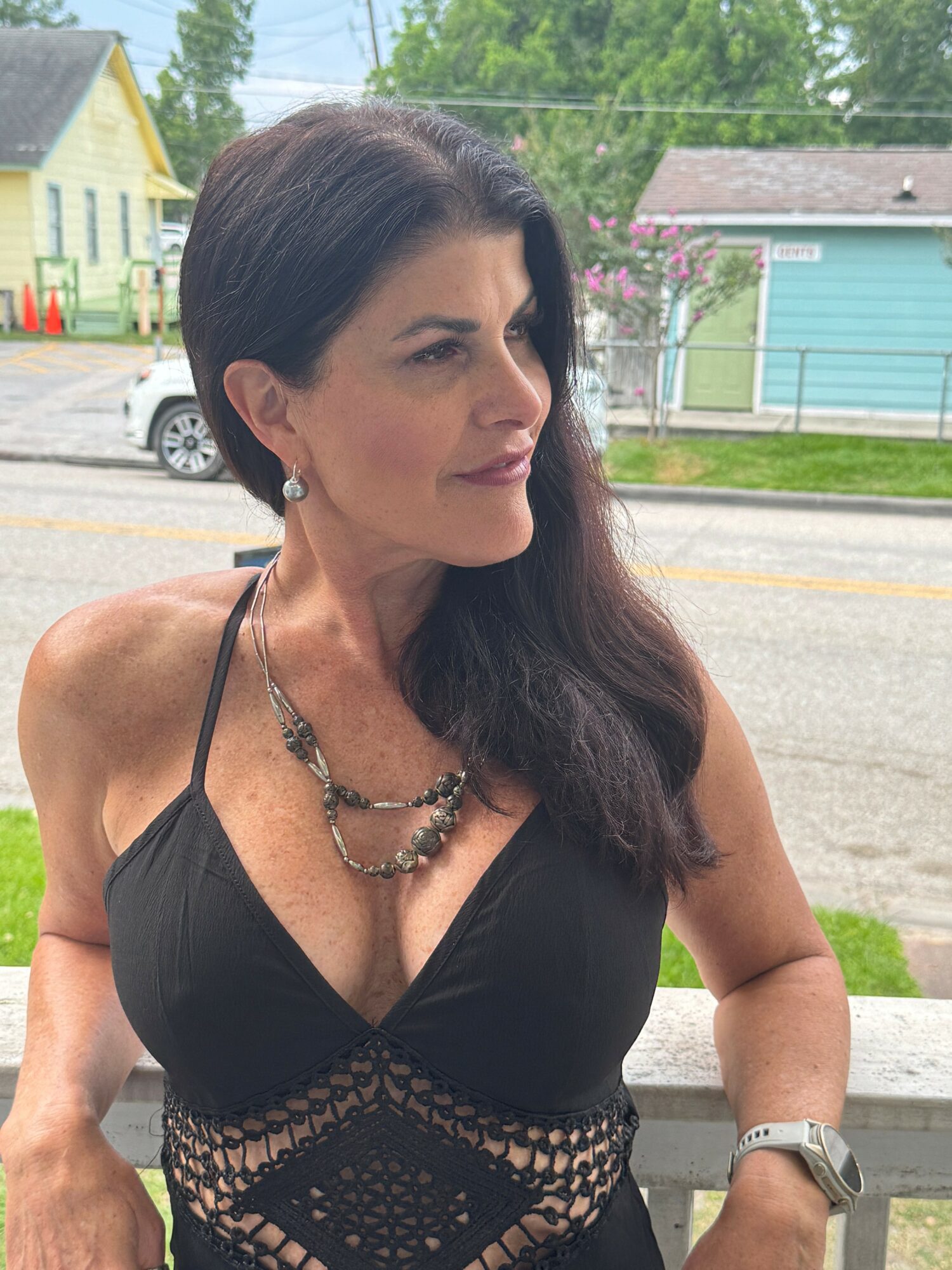 Woman with dark hair in a black dress outdoors, wearing jewelry and a watch, with houses and trees in background.