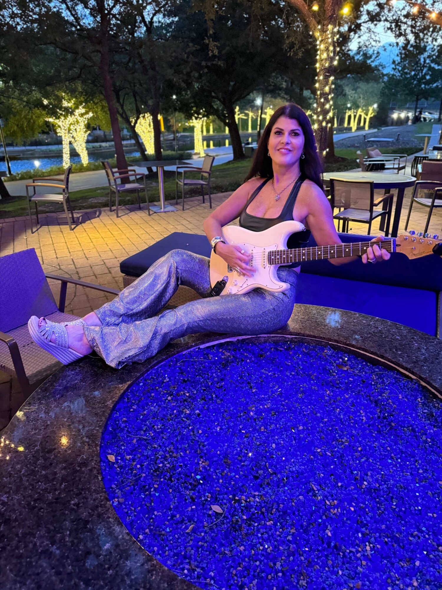 Woman sitting outdoors with a guitar, illuminated trees and string lights in the background, sitting on a blue couch.