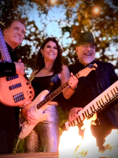 Three musicians playing guitars and keyboard outdoors with trees and sky in background, smiling and performing.