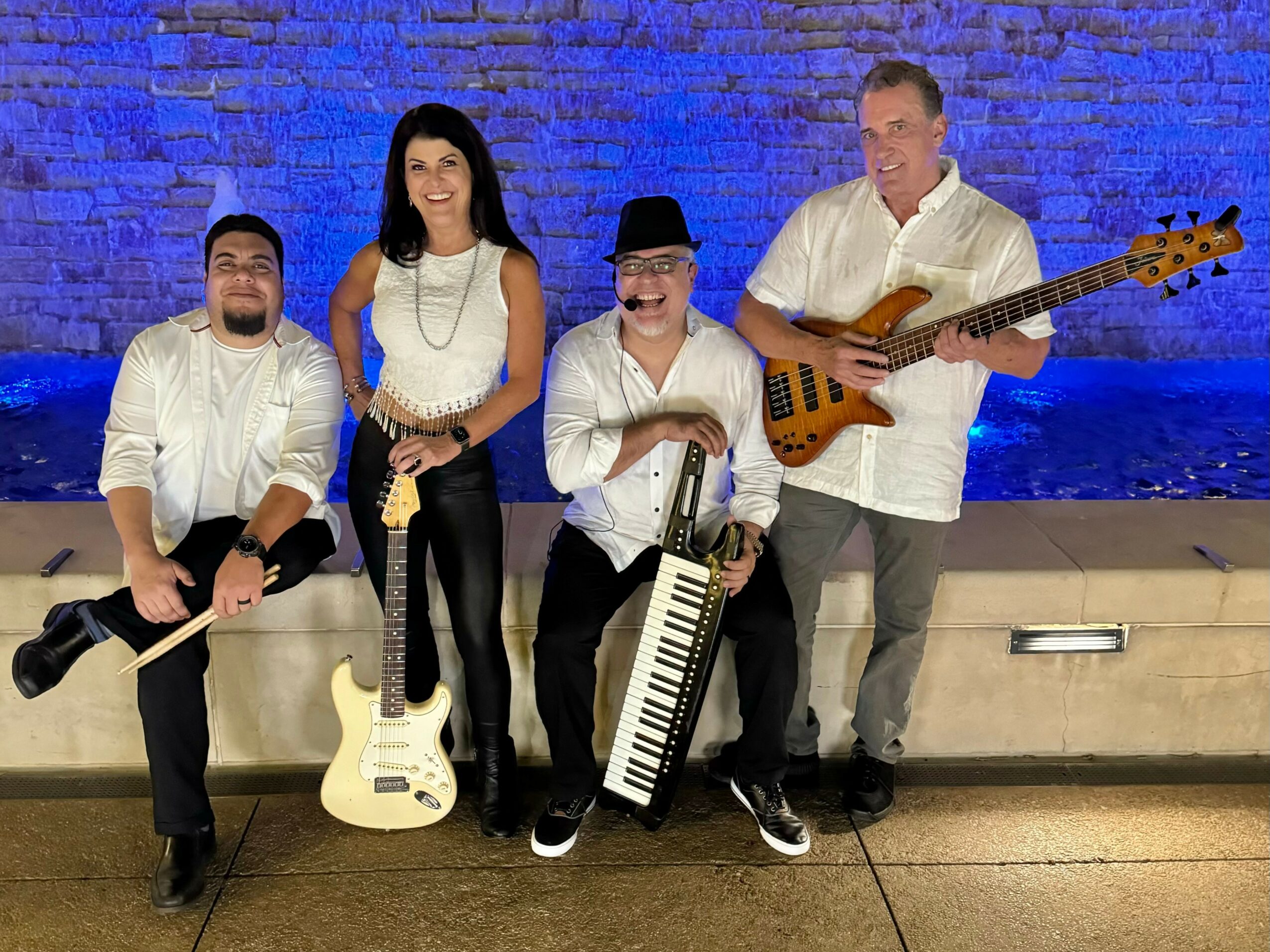 Four musicians pose with instruments against a blue-lit brick wall, smiling and dressed in white shirts and dark pants.