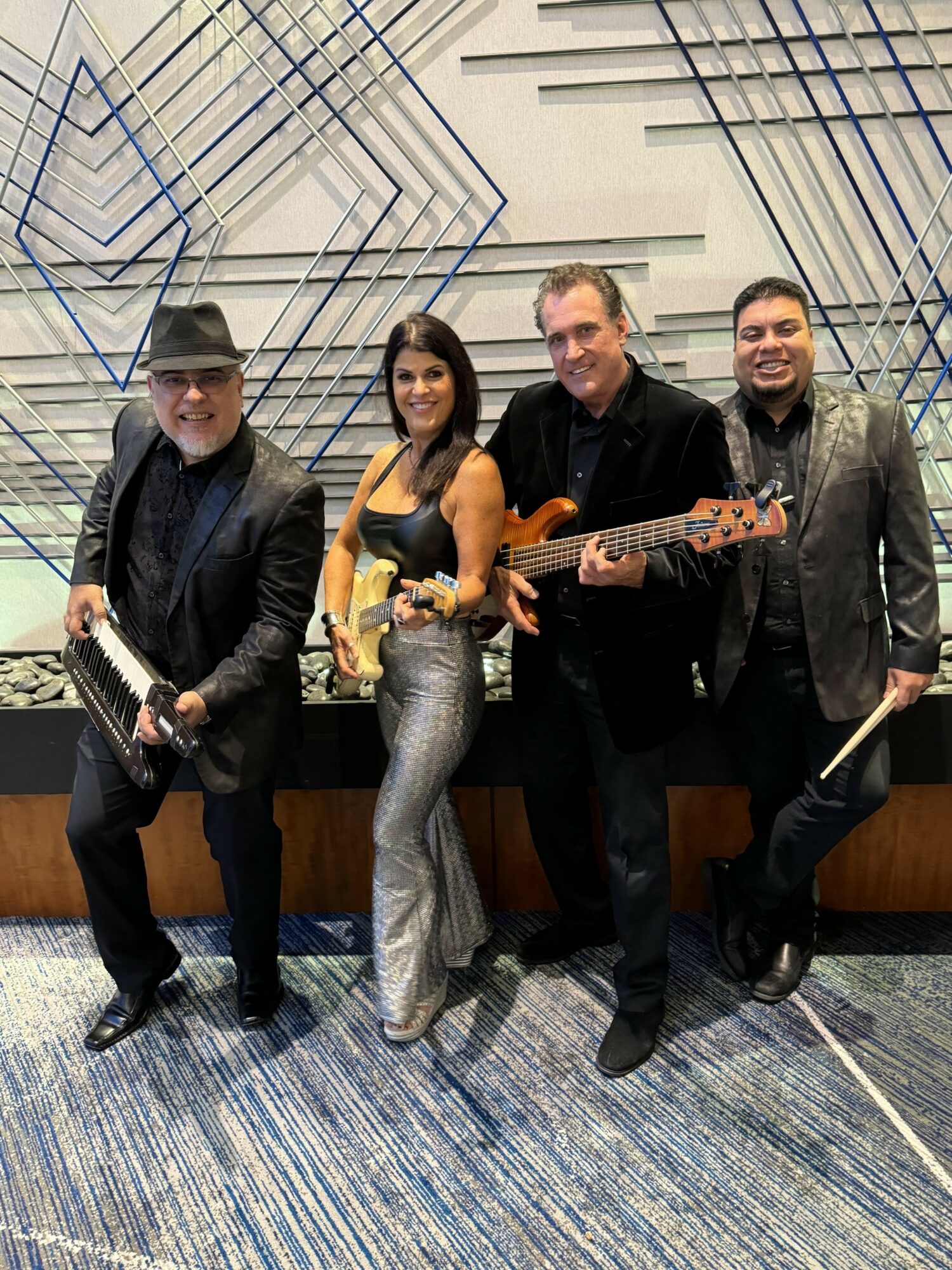 Four musicians pose with instruments in a modern indoor setting, smiling at the camera.
