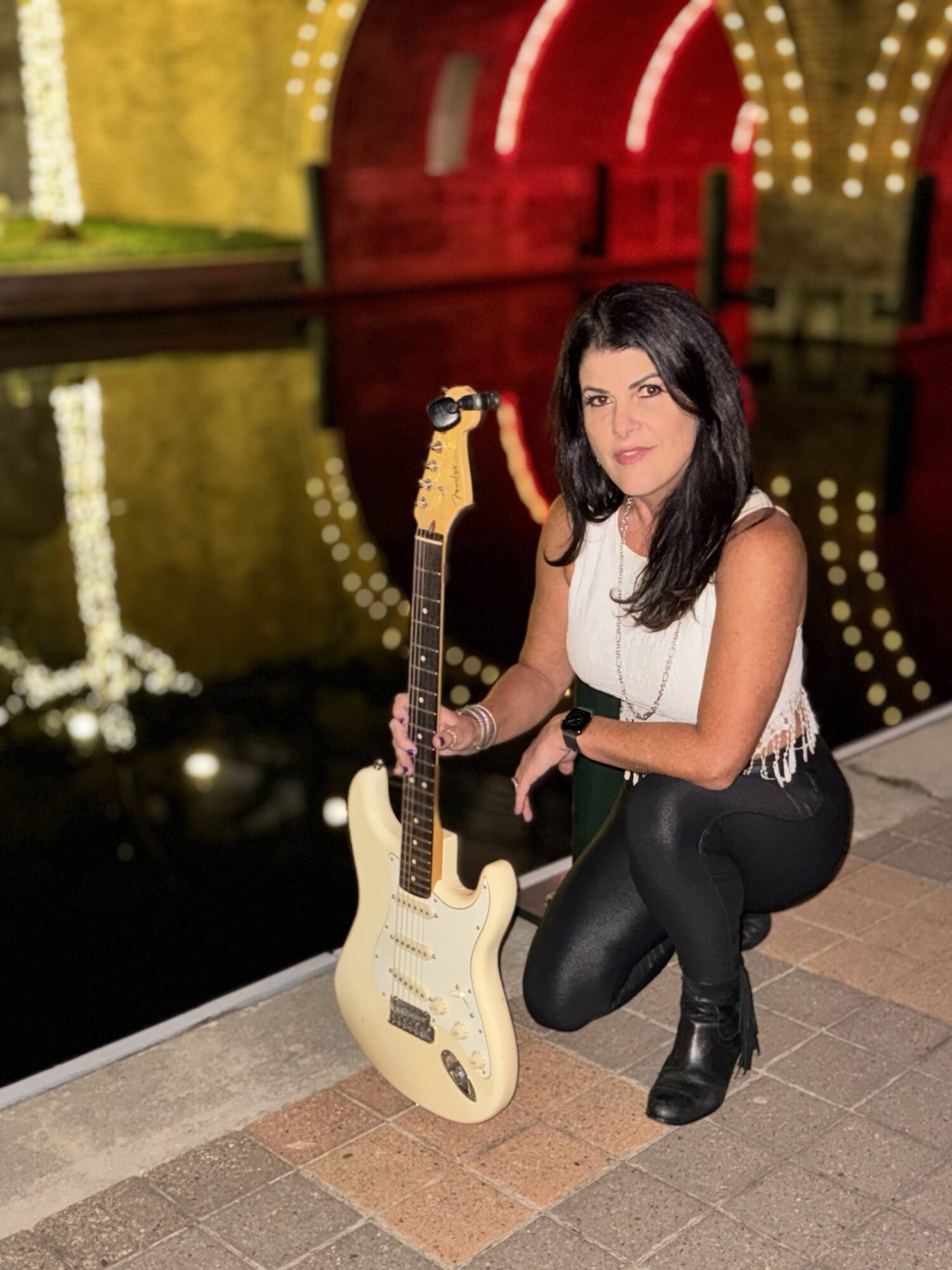 Woman crouching next to an electric guitar near a body of water with colorful lights reflected, nighttime scene.