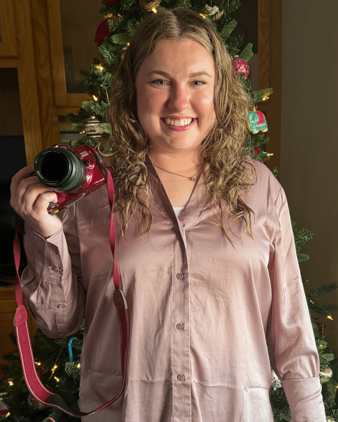 Young woman with curly hair smiling, holding a red camera, standing in front of a decorated Christmas tree.