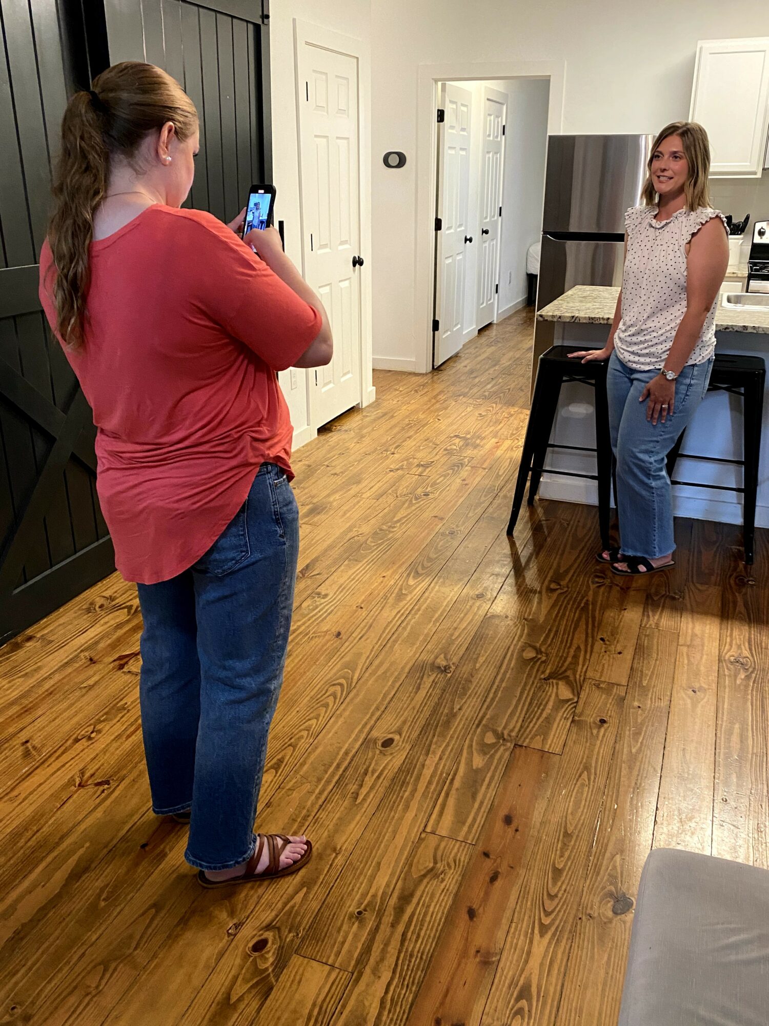 Two women in a kitchen, one taking a photo of the other, with wooden floors and white walls.