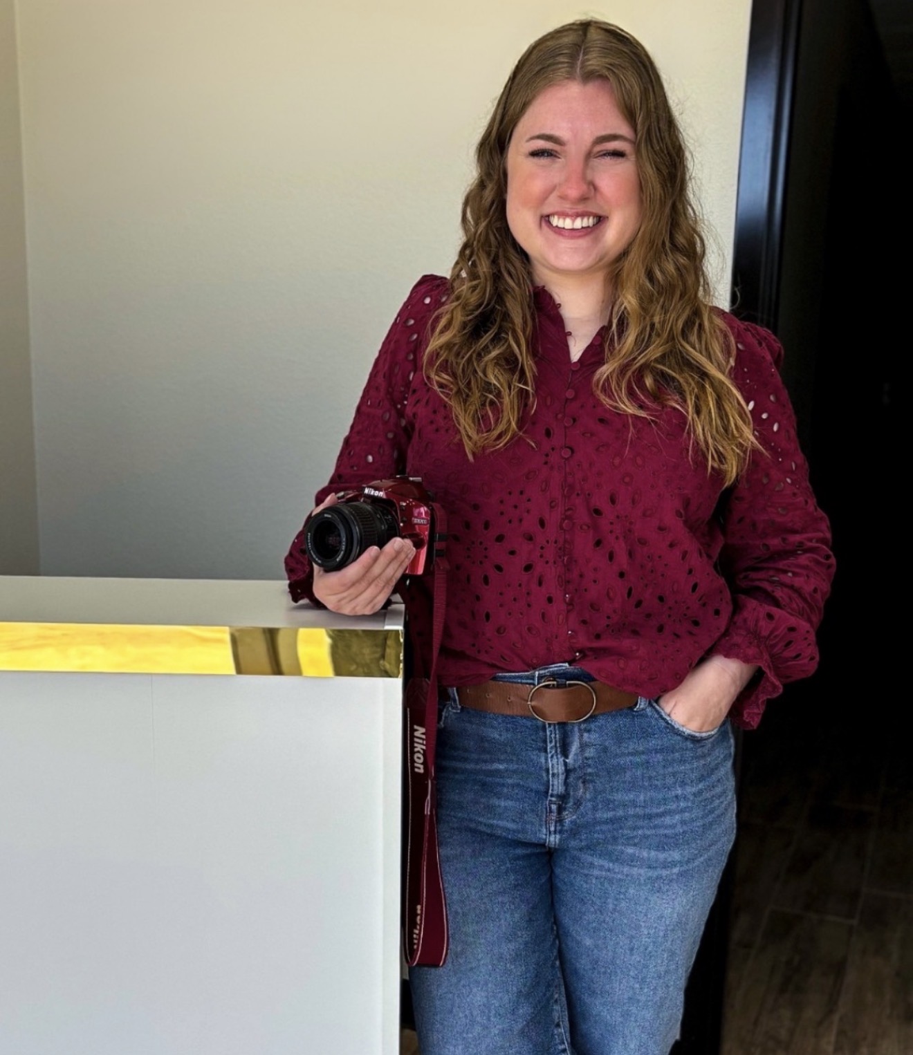 Young woman with long wavy hair smiling, holding a camera, wearing a maroon sweater and blue jeans, standing indoors.