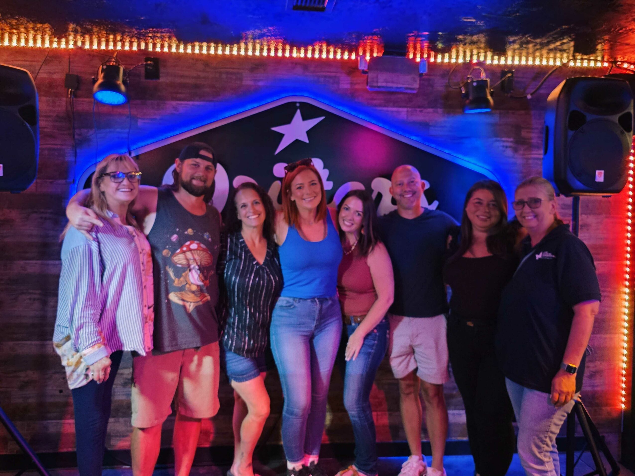 Group of seven people standing together in front of a decorated background with a star, illuminated by colorful lights.