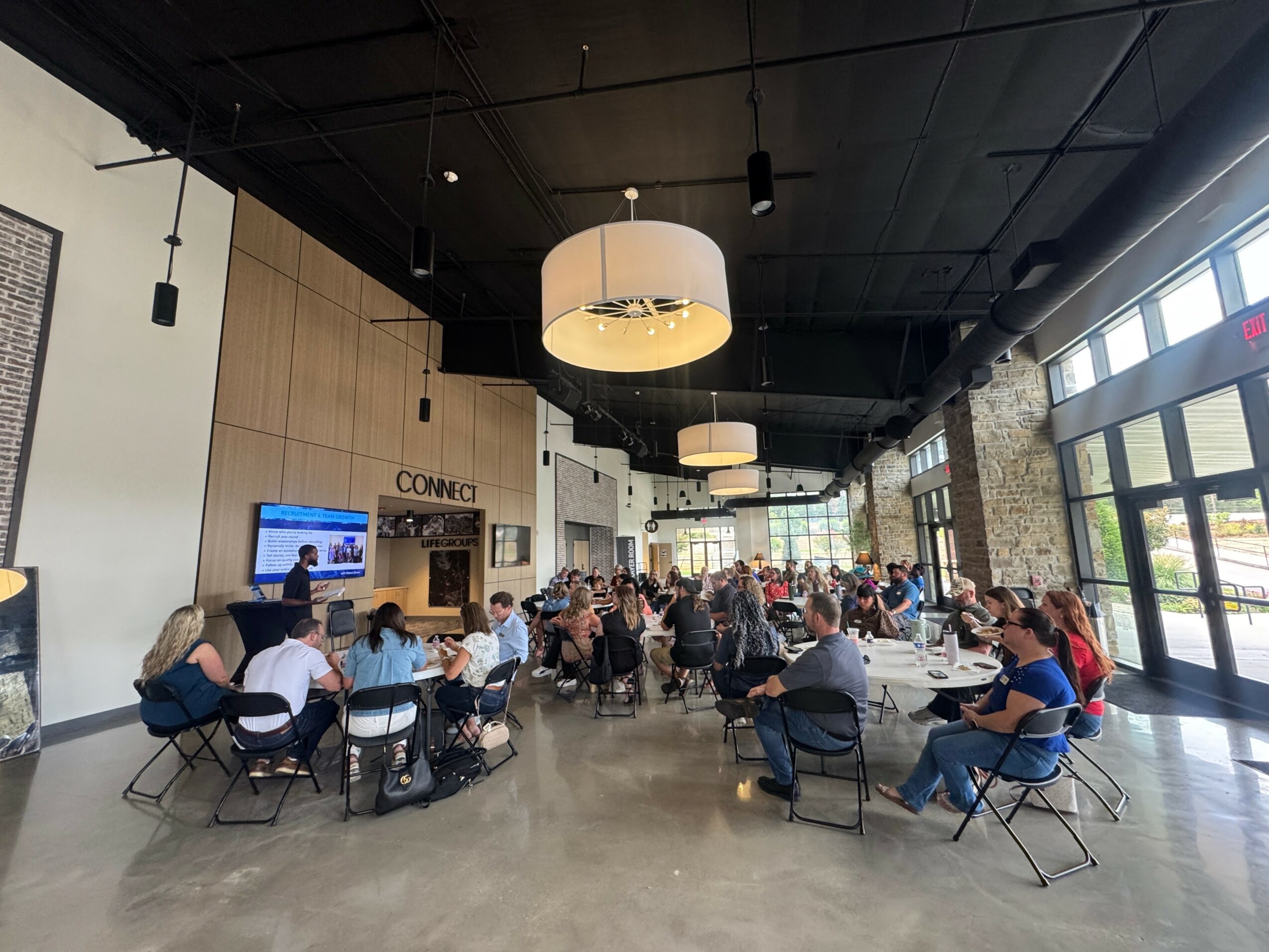 People sitting at tables in a spacious, modern indoor space with large windows and hanging lights.