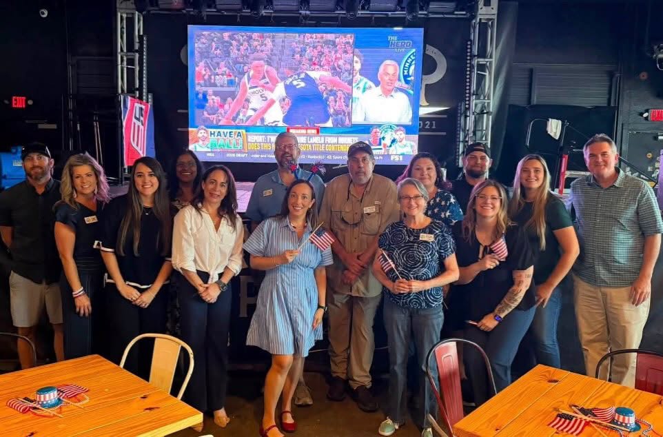 Group of people standing together in a room with a large screen showing a sports event in the background.