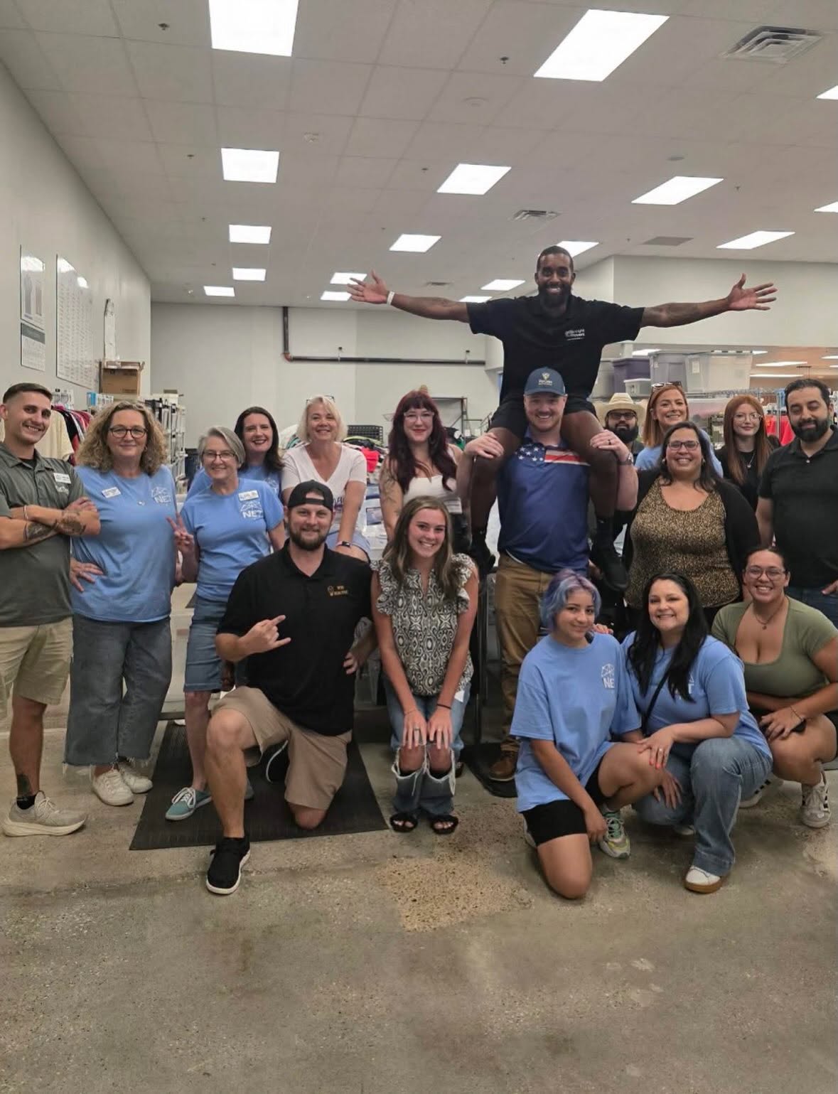Group of people in a room, some wearing blue shirts, with one person standing on others' shoulders, arms outstretched.