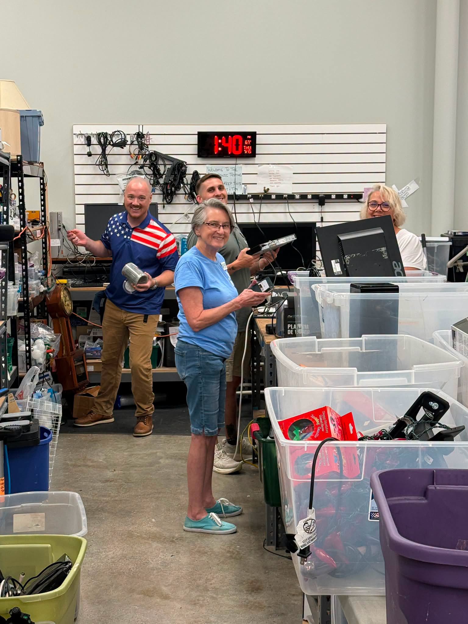 Four people in a store checkout area, with shelves and containers around, smiling and holding items, digital clock showing 1:40.