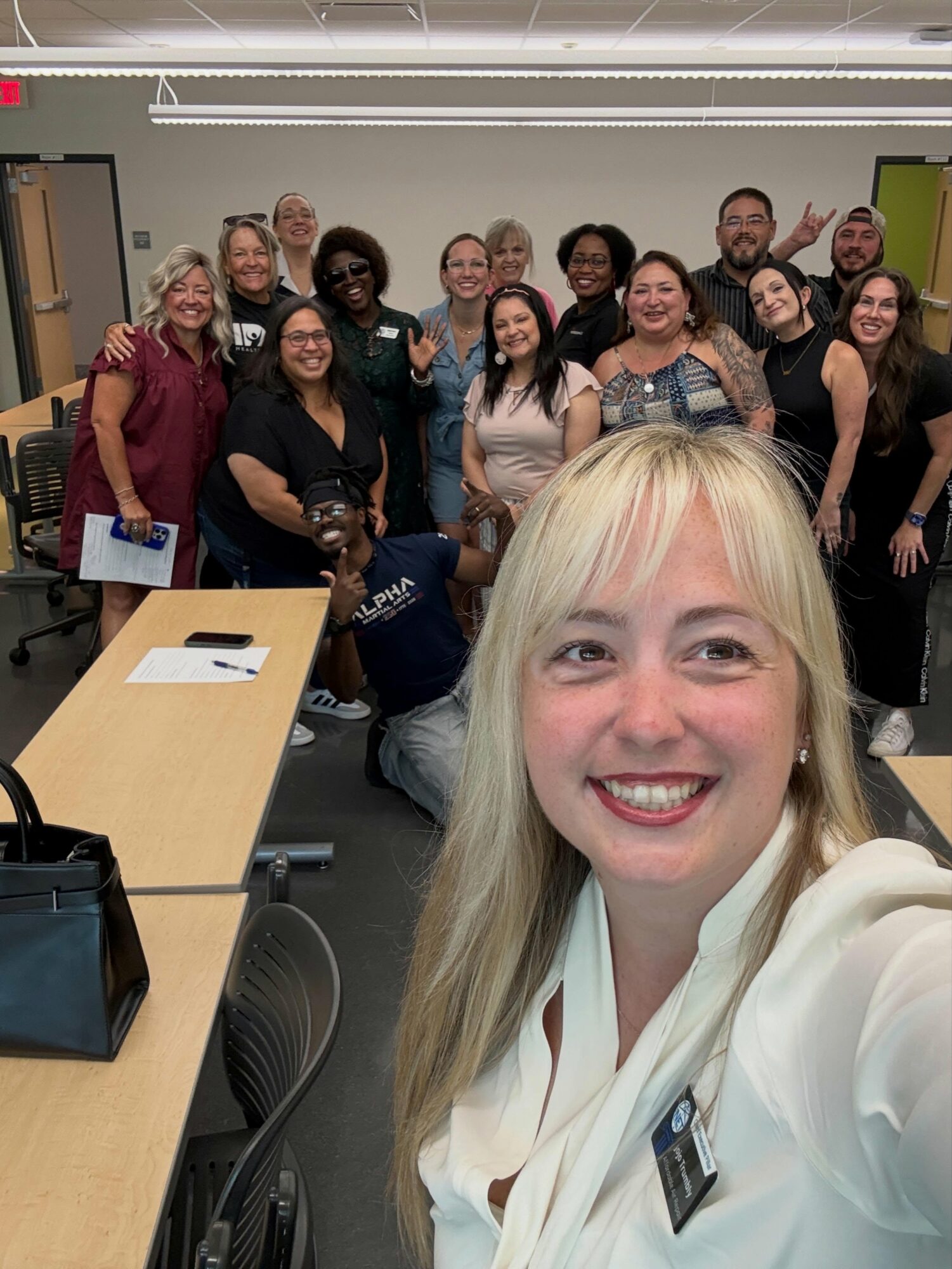 Group of diverse people smiling in a classroom, with a woman taking a selfie in the foreground.