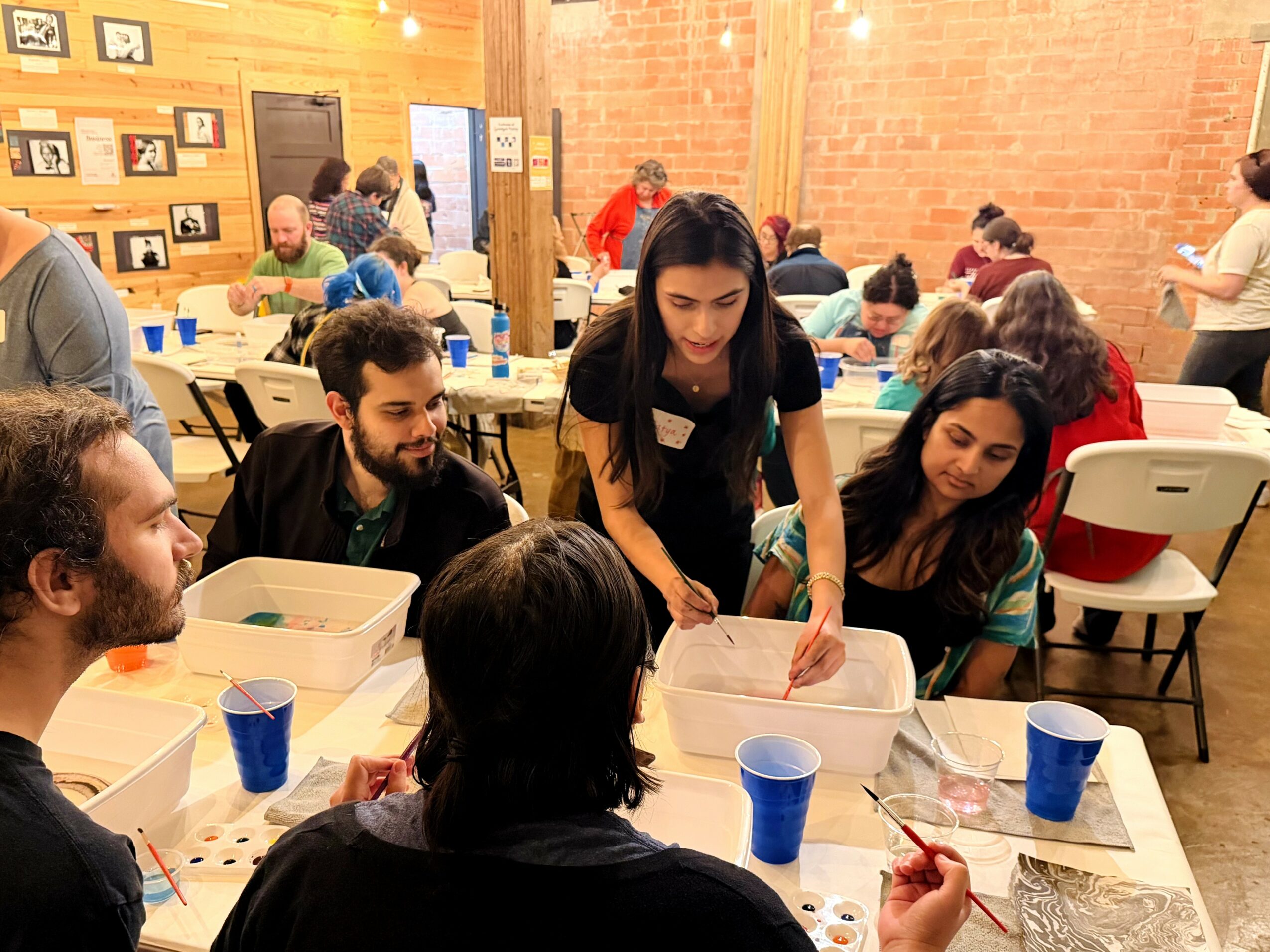 Group of people sitting at a table with art supplies, woman standing and demonstrating, others watching attentively, indoor setting with brick wall.