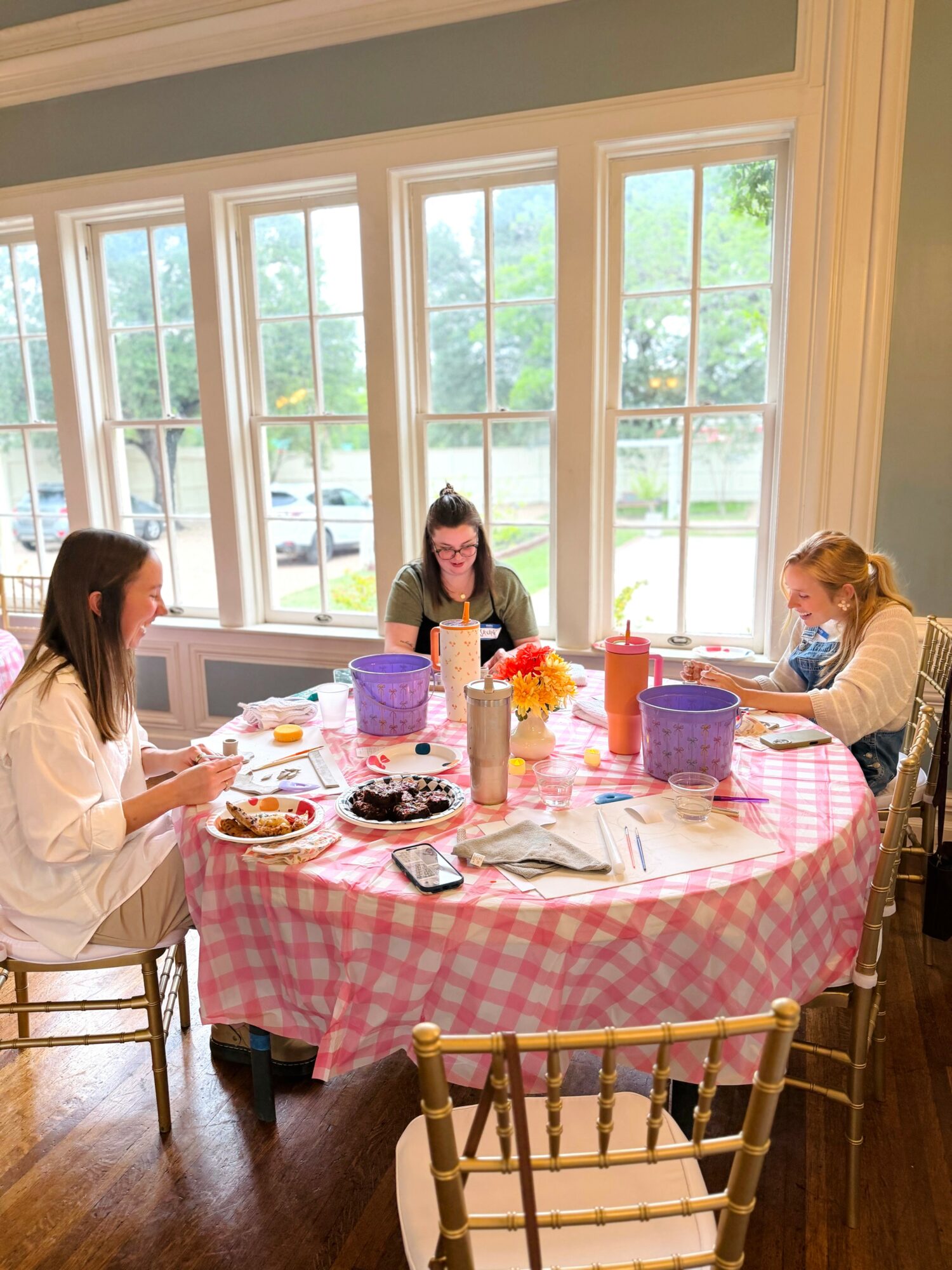 Three women sit around a round table with a pink checkered tablecloth, enjoying a meal in a bright room with large windows.
